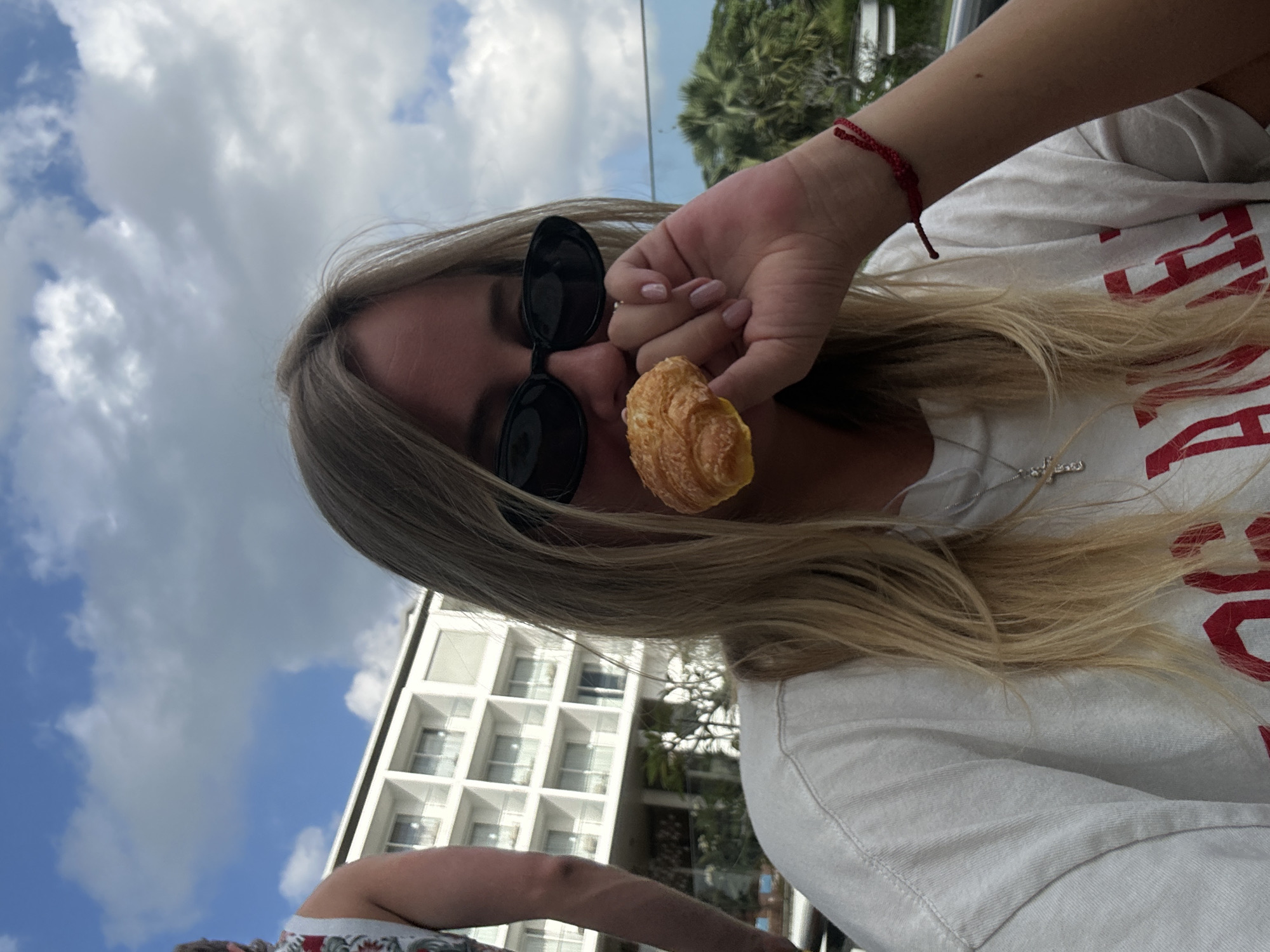 A young woman with blonde hair and sunglasses is in the foreground, holding a croissant to her mouth. She is wearing a white t-shirt with red lettering and a red bracelet on her left wrist. In the background, a modern white building with balconies is visible, along with palm trees and a blue sky with clouds. The scene appears to be outdoors, likely at a resort or hotel in Kuta Selatan, Indonesia, given the architectural style and vegetation.