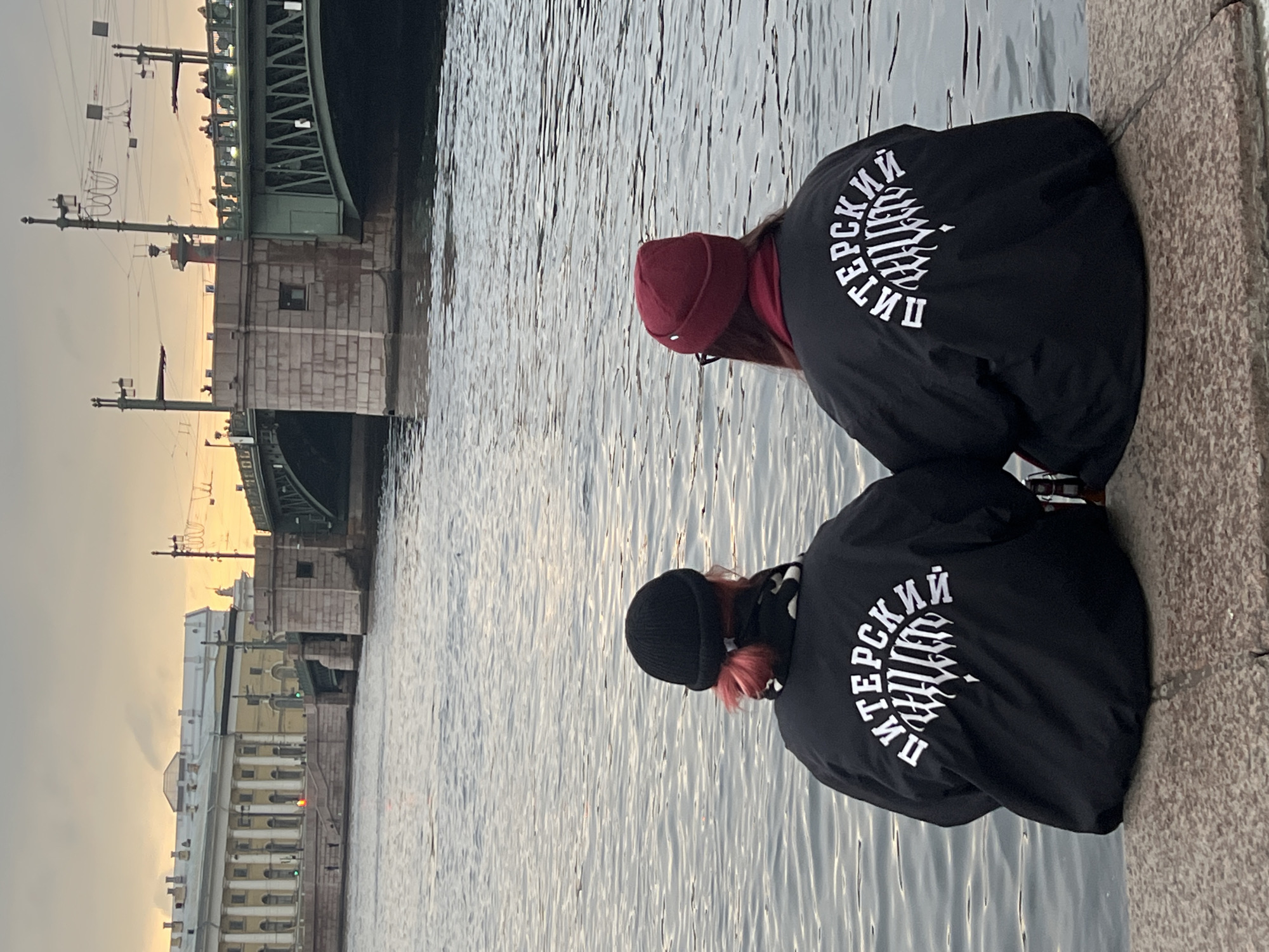 Two individuals are sitting on a concrete bank overlooking a body of water, likely a river or canal. Both are wearing black jackets with white lettering in Russian, possibly "Питерский стиль" or "Petersburg style," and dark beanies. One is wearing a burgundy beanie, while the other is wearing a black beanie with some pink hair visible.
In the background, there is a cityscape that includes a bridge, buildings, and overhead wires. The lighting suggests it is either sunrise or sunset. The location is likely in Saint Petersburg, Russia, as indicated by the Russian lettering on the jackets and the architecture of the buildings.
