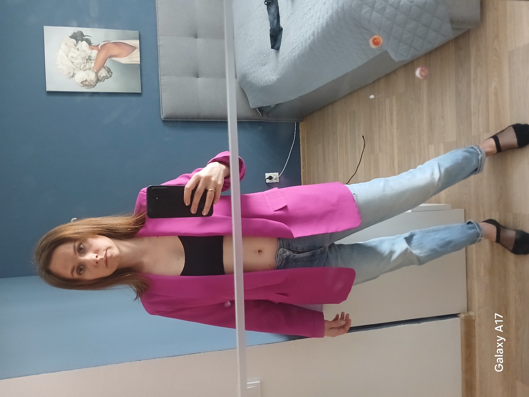 A woman is posing for a selfie in what appears to be her apartment. She is wearing a black crop top, light blue jeans, a bright pink blazer, and black mesh socks. She is holding a black smartphone, presumably to take the picture. The background shows a blue wall with a framed picture, a gray headboard, and a bed with a light blue bedspread. The floor is made of light-colored wood. The location is in a residential setting, though without additional context no assertion on its presence in Yekaterinburg, Russia can be asserted. The image also contains the brand name "Galaxy A17" written in the bottom right corner.