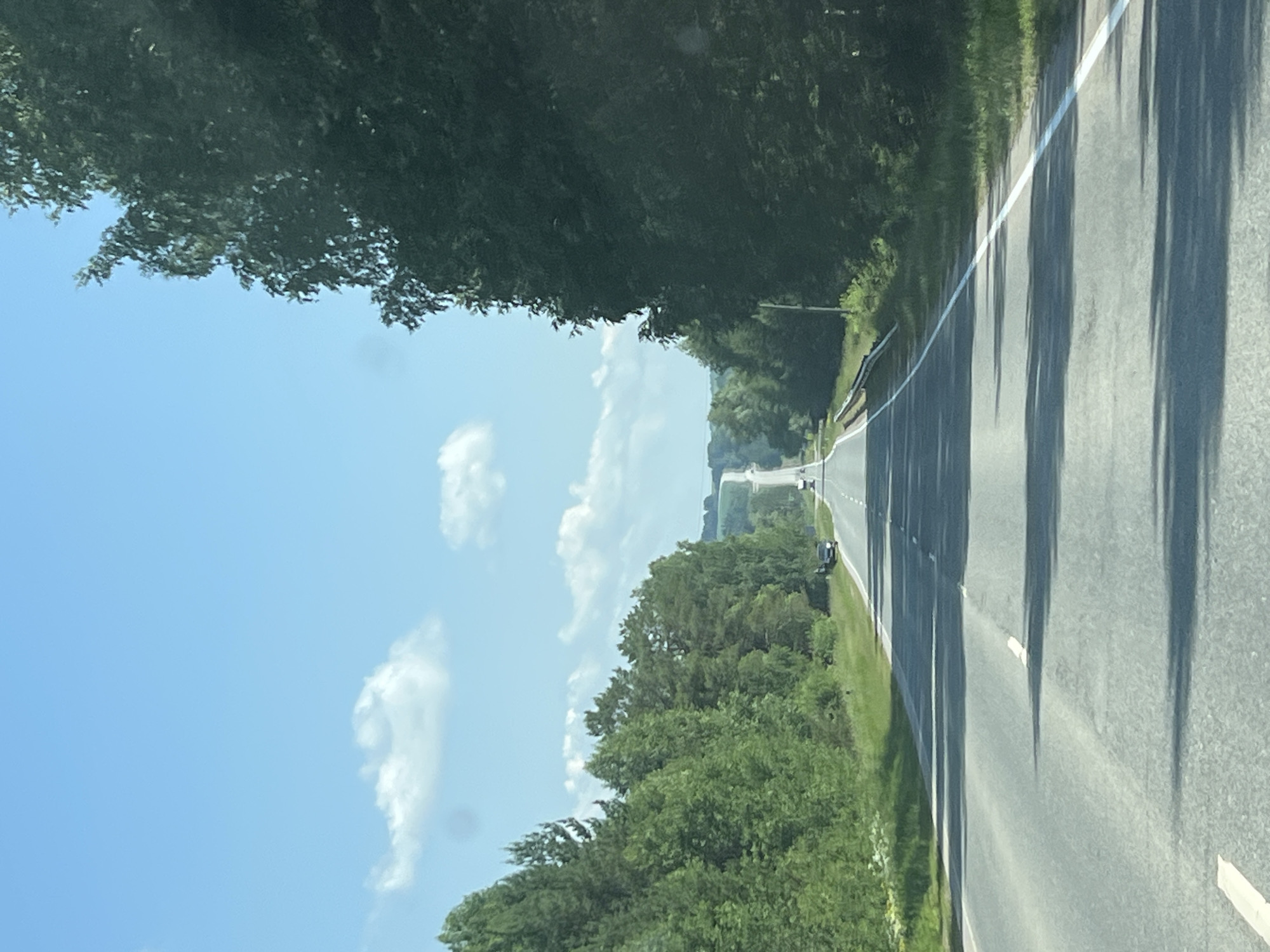 The image shows a long, straight asphalt road stretching into the distance, lined by dense green trees on both sides. The sky is bright blue with a few scattered white clouds. The road has white lane markings. In the distance, the road appears to ascend a gentle hill, and there are a few vehicles visible on the road, including a black car parked on the right shoulder and what appears to be a truck in the distance. The overall scene depicts a scenic drive on a clear day through a natural, possibly rural, environment. The image does not provide any specific visual information that would indicate it is located in the city of Kirillovka, Russia.