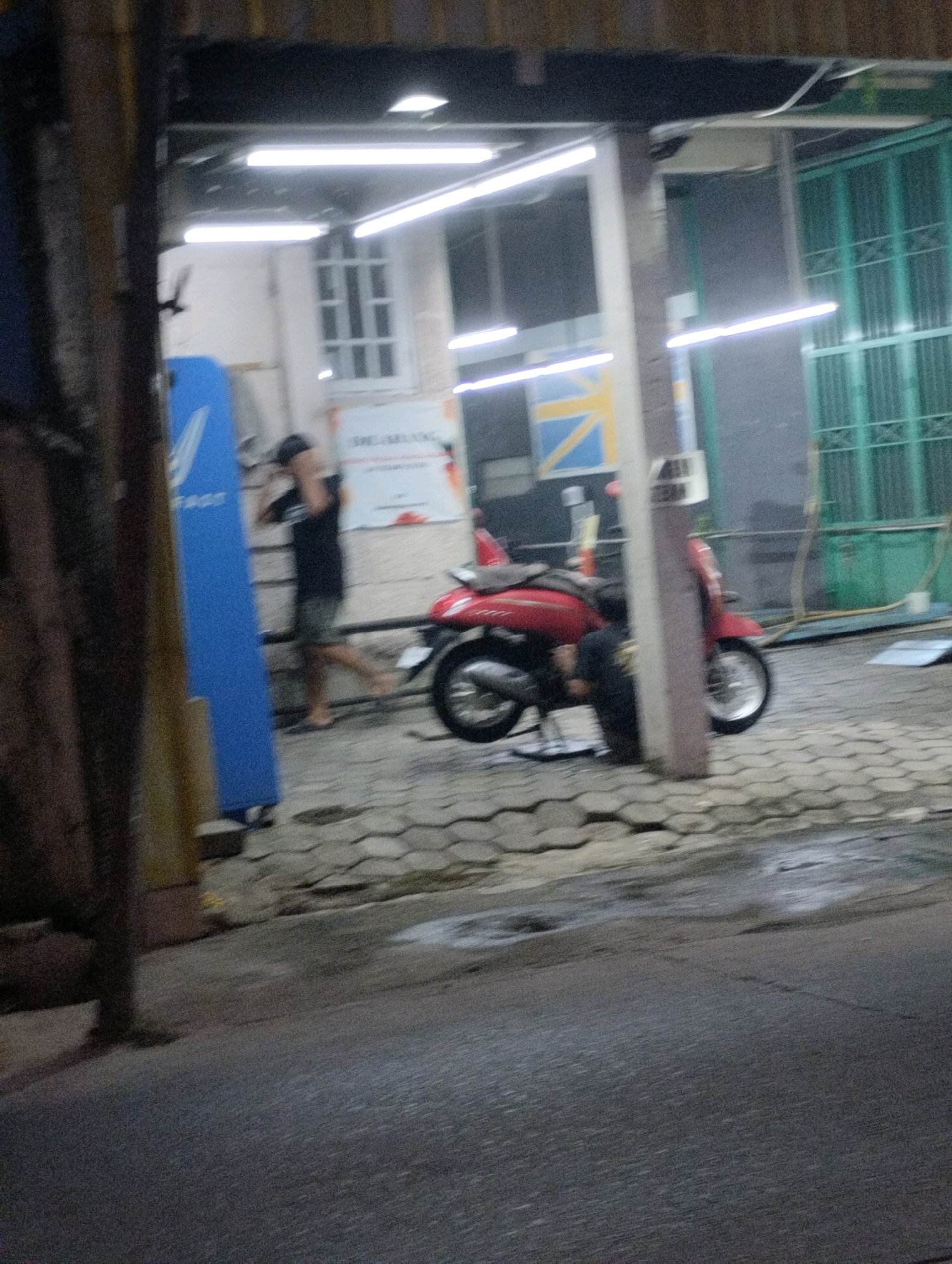 The image shows a dimly lit outdoor scene, likely at night, in what appears to be an urban or commercial area. A person, blurred due to motion, is visible in the mid-ground, wearing a dark t-shirt and shorts, and a cap. They are walking away from the viewer towards a red scooter parked on a patterned tiled surface. The scooter has a cover draped over its rear. To the left of the person, a tall, bright blue object with white lettering is standing upright. In the background, fluorescent lights illuminate the area, highlighting a building facade with a window and a large green gate with vertical slats. The ground in front of the building is wet, suggesting recent rain or cleaning. The overall impression is of a street or a service area, possibly a shop or a vehicle repair establishment in Bekasi, Indonesia, given the context.