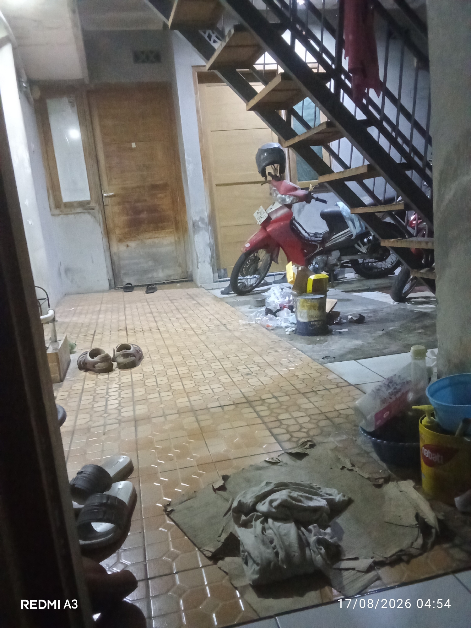 The image depicts an interior space, likely a common area or hallway, with a red scooter parked under a metal staircase. The floor is tiled with a beige hexagonal pattern. To the left, there is a wooden door with a glass pane. In the foreground, on the floor, are two pairs of sandals. Further back, near the scooter, there are several containers and scattered debris, including plastic bags and a yellow bucket with the word "Nabati" on it. A pile of clothing is also visible on a piece of cardboard in the lower right portion of the image. The lighting suggests it is either dusk or night, with artificial light sources illuminating the scene. There are no people visible in the image. The timestamp in the lower right corner reads "17/08/2026 04:54". The location is identified as Banaran, Indonesia.