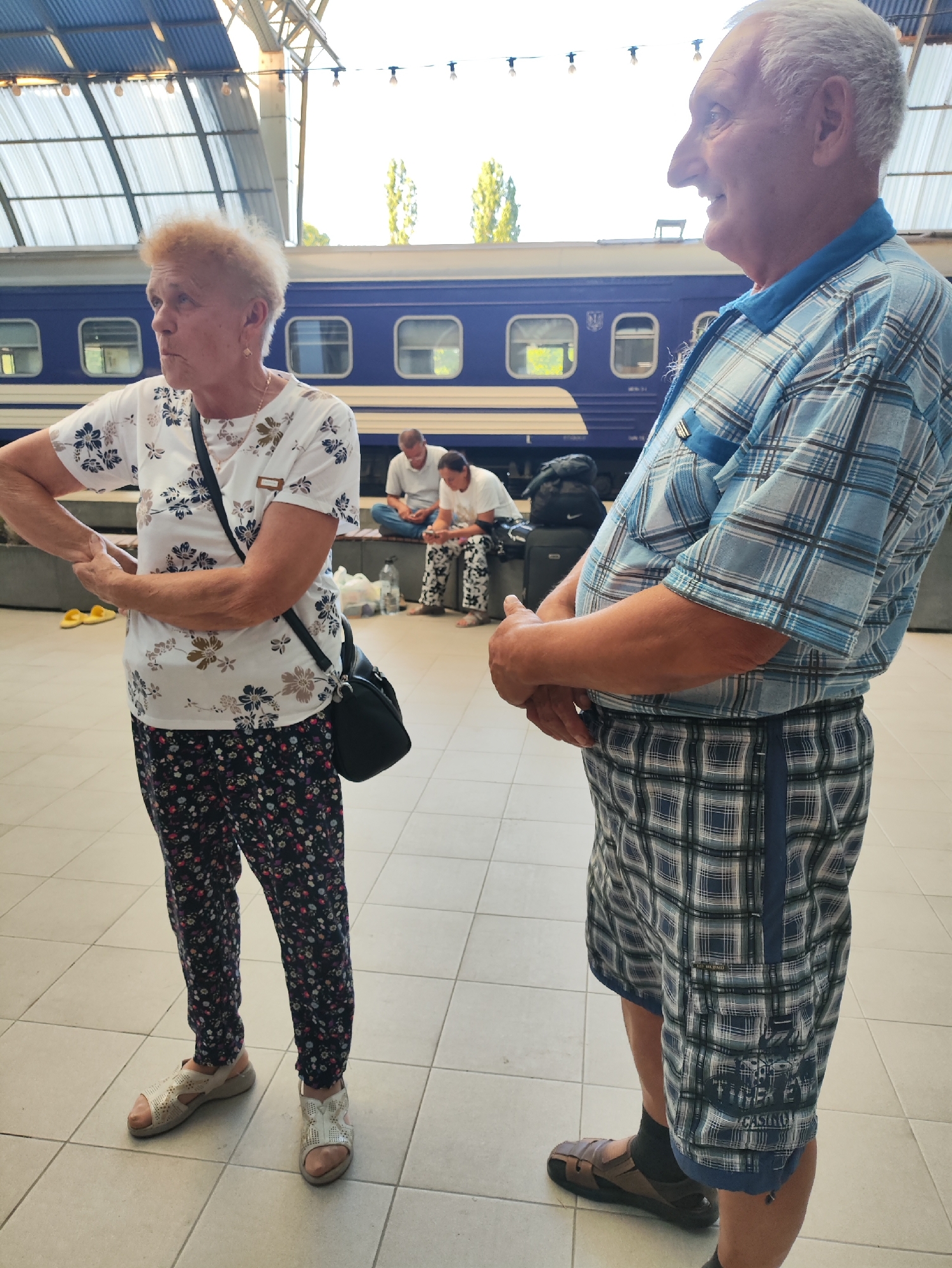 An elderly woman with short, curly blonde hair stands on a tiled platform, facing left and looking away. She wears a white t-shirt with a floral pattern and dark pants with a similar print. A black crossbody bag is slung over her shoulder. Her hands are clasped in front of her. In the background, two men sit on a bench near a blue train. One man wears a white polo shirt and jeans, looking at his phone. The other, in a white t-shirt, appears to be looking down. Luggage is visible near them. To the right of the woman, an elderly man with gray hair, wearing a blue plaid shirt and plaid shorts, stands facing right. His hands are clasped in front of him. The scene appears to be at a train station, possibly in Chișinău, Moldova, with a covered platform overhead and trees visible in the distance.