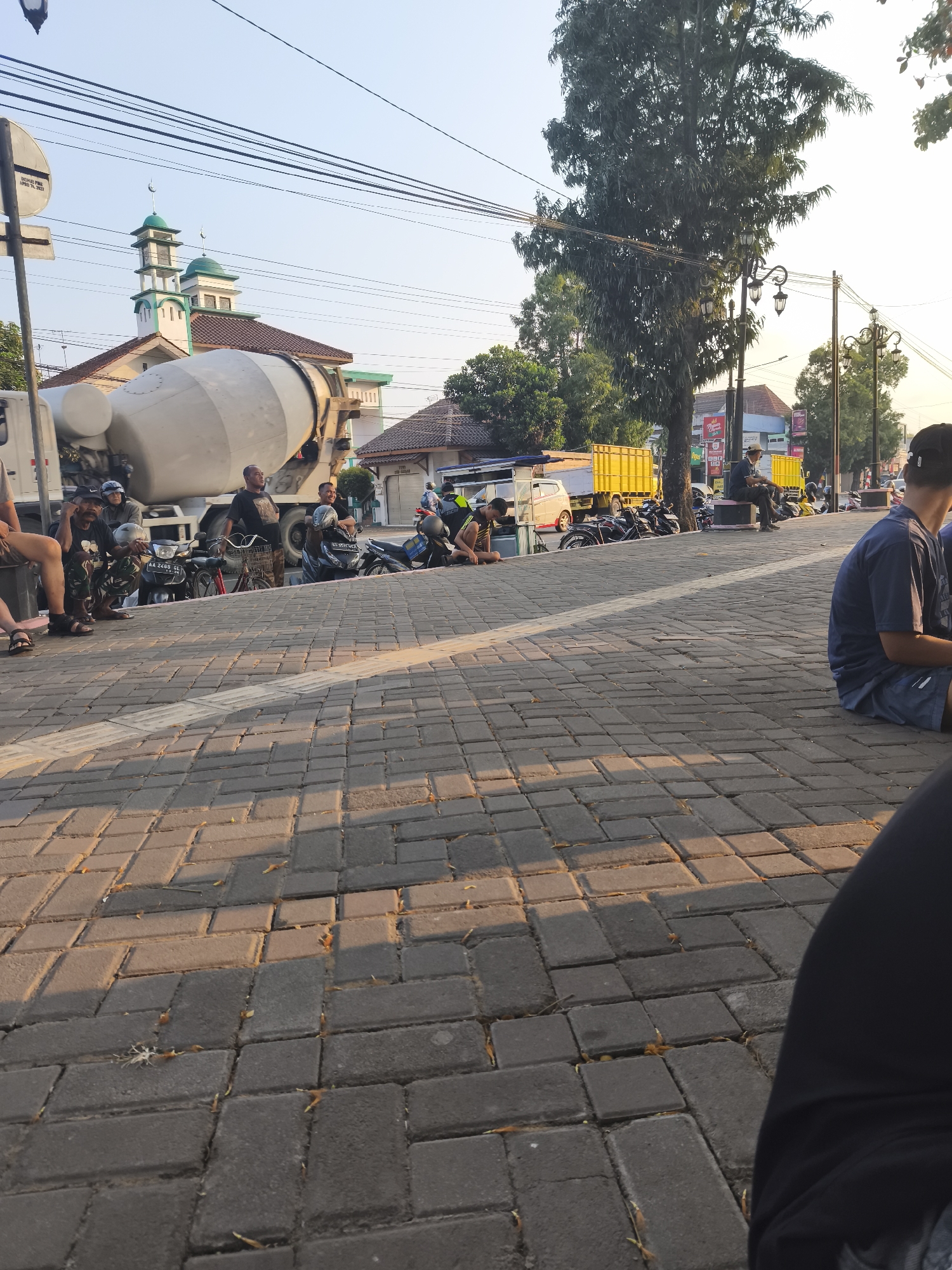 The image captures a street scene in Kutoarjo, Indonesia, likely near a mosque, as evidenced by the distinctive green-domed structure in the background. A large concrete mixer truck is parked prominently on the left side of the frame, its drum facing towards the viewer. Several individuals are seated on the ground and on motorcycles, suggesting a temporary pause or gathering. The foreground is dominated by a paved walkway constructed from interlocking gray bricks, casting long shadows due to the low angle of the sun. In the mid-ground, more motorcycles are parked, and a yellow truck is visible. The background reveals buildings, trees, and utility poles with overhead wires, typical of an urban environment. The sky is clear and bright, indicating daytime.