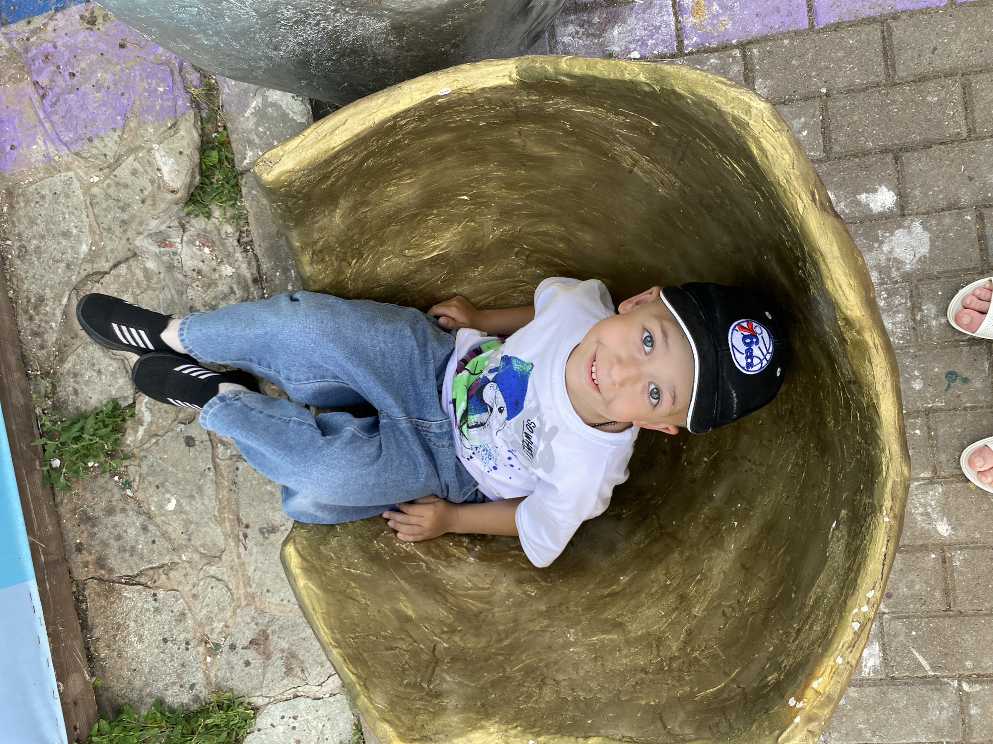 A young boy is lying on his back inside a large, golden, bowl-shaped sculpture. He is wearing a white t-shirt with a graphic print, blue jeans, black sneakers with white stripes, and a black baseball cap with a logo. The sculpture is situated outdoors on a paved area with visible cracks and patches of grass. In the background, there are large stone slabs and sections of purple-painted pavement. The scene appears to be in a public space, possibly a park or an outdoor art installation. The provided crops suggest the location is in the city of Izhevsk, Russia.