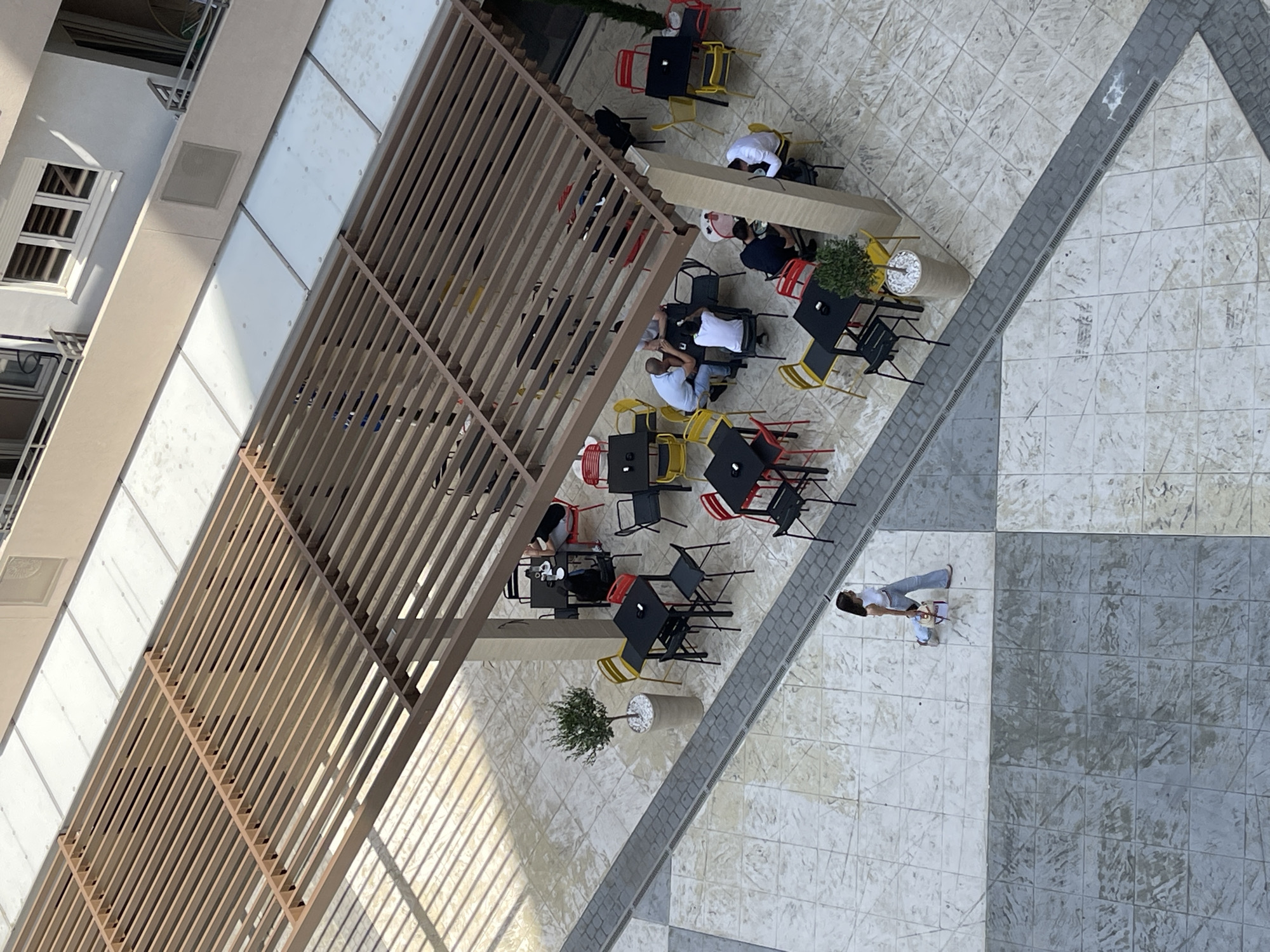 An overhead view captures an outdoor seating area of a cafe or restaurant. A wooden pergola covers a portion of the seating arrangement, casting striped shadows on the tiled ground. Several black tables with red and yellow chairs are occupied by patrons. One table has two individuals, another has three, and two tables have single individuals. A woman in blue jeans and a white top walks across the tiled area on the lower left, pulling a small red and white wheeled suitcase. A potted tree sits to the left of the seating area. The ground is tiled with a pattern of lighter and darker square tiles, with a drainage grate running along a diagonal path. In the background, a building with balconies and windows is visible. The scene appears to be in Podgorica, Montenegro, given the architectural style and outdoor cafe culture.