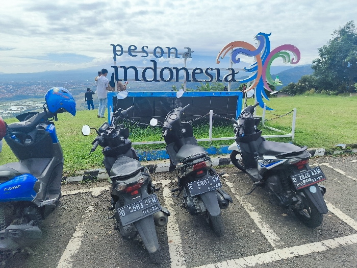 Four motorcycles are parked in a parking lot in Majalengka, Indonesia, located at latitude -6.846 and longitude 108.204. The motorcycles are parked in a row, facing the same direction. A sign that says "Pesona Indonesia" is in the background, with a bird-like sculpture. The sign is adorned with colorful, stylized swirls and an inscription that reads “pesona Indonesia”. The parking lot is paved with white stripes separating the parking spaces. The parking lot is surrounded by a grassy area. The sky is cloudy. The image is likely captured by a person standing near the motorcycles.