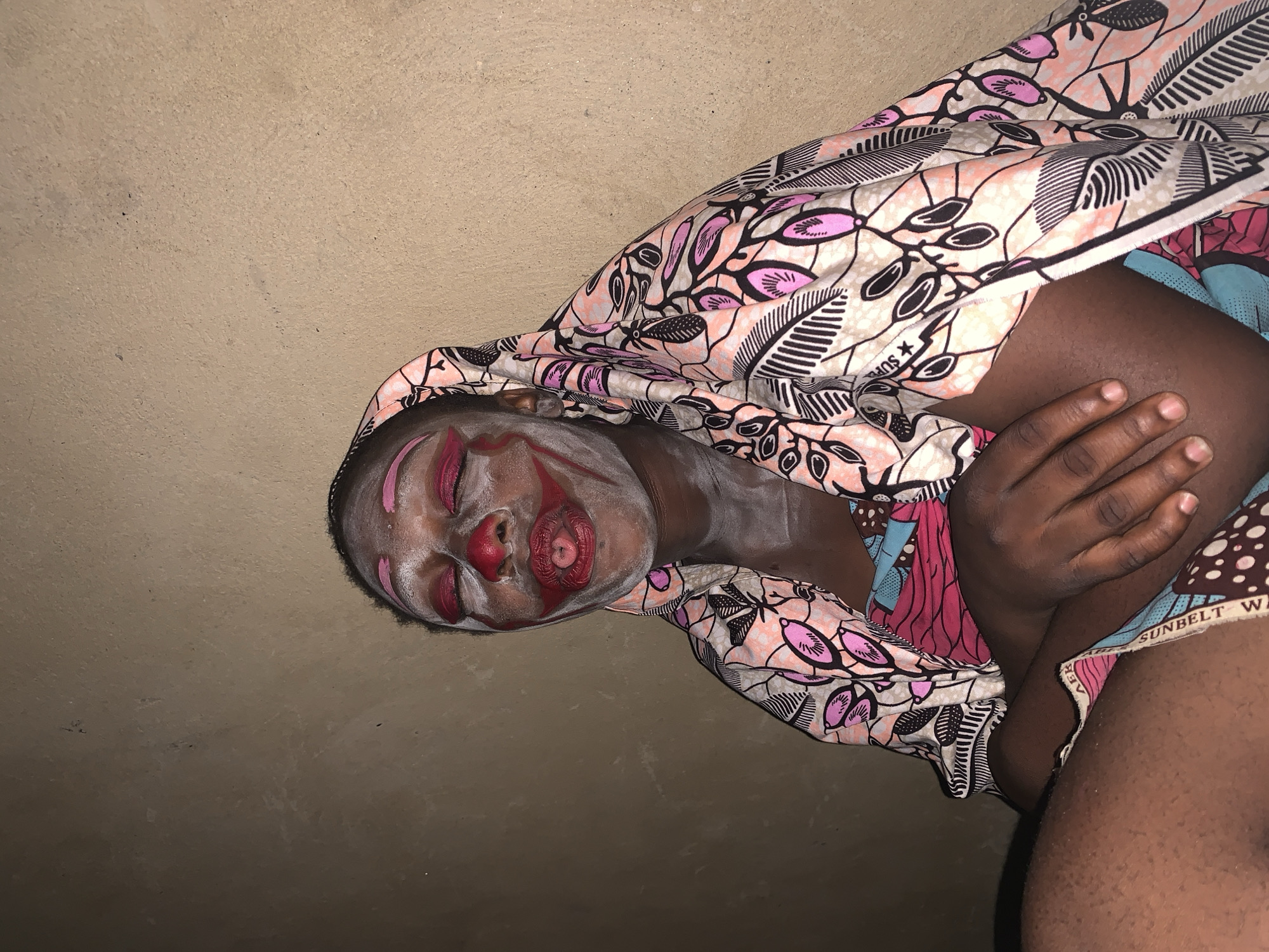 A close-up, medium shot captures a person with their eyes closed, face painted as a clown, and a colorful patterned headscarf and clothing. The person is sitting, with their hands resting on their lap. The background is a textured, light brown wall. The lighting appears to be artificial, casting a slight shadow. The person's facial makeup includes white and red paint, with exaggerated features resembling a clown's. Their tongue is sticking out, and a red substance, likely lipstick, is smeared around the mouth. The headscarf features a pink and black floral pattern. The person is wearing a blue and pink patterned garment underneath. The scene is set indoors, with the wall suggesting an interior setting, potentially a home or a community space in Agban, Nigeria.