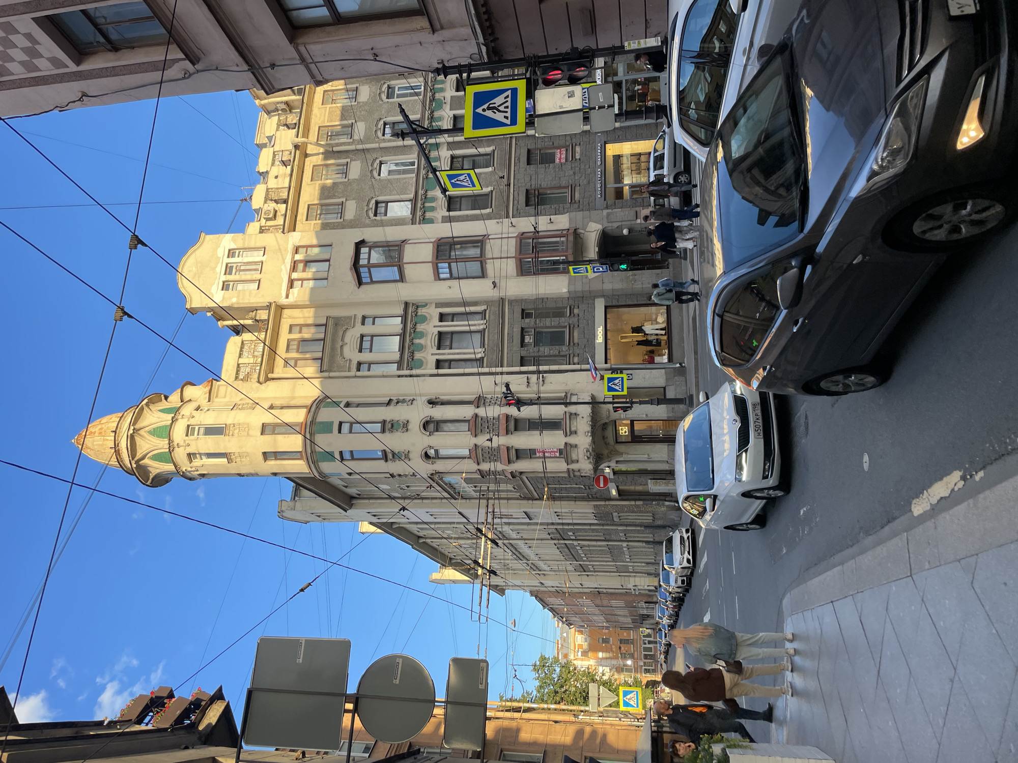 The image captures a street scene in Saint Petersburg, Russia, on a clear day with a blue sky and golden sunlight illuminating the buildings. The architecture is diverse, featuring a prominent art nouveau tower with a decorative dome and green accents, alongside apartment buildings with classical and baroque elements, characterized by bay windows and ornate facades.
The street is paved with asphalt and lined with sidewalks. Numerous cars are parked along the right side of the street, and several are in motion, including a white Skoda Rapid and a dark gray Hyundai Solaris in the foreground. Overhead, a complex network of electrical wires, likely for public transport, crisscrosses the sky.
Several pedestrians are visible on the sidewalks. On the left, two women are standing and looking at their phones. On the right sidewalk, a group of people are walking. Traffic signals and pedestrian crossing signs are present, indicating an urban intersection.
Businesses are visible at street level, with storefronts labeled with names like "IDOL" and what appears to be a confectionery shop. The overall impression is a vibrant, historic urban environment with active street life and traffic.