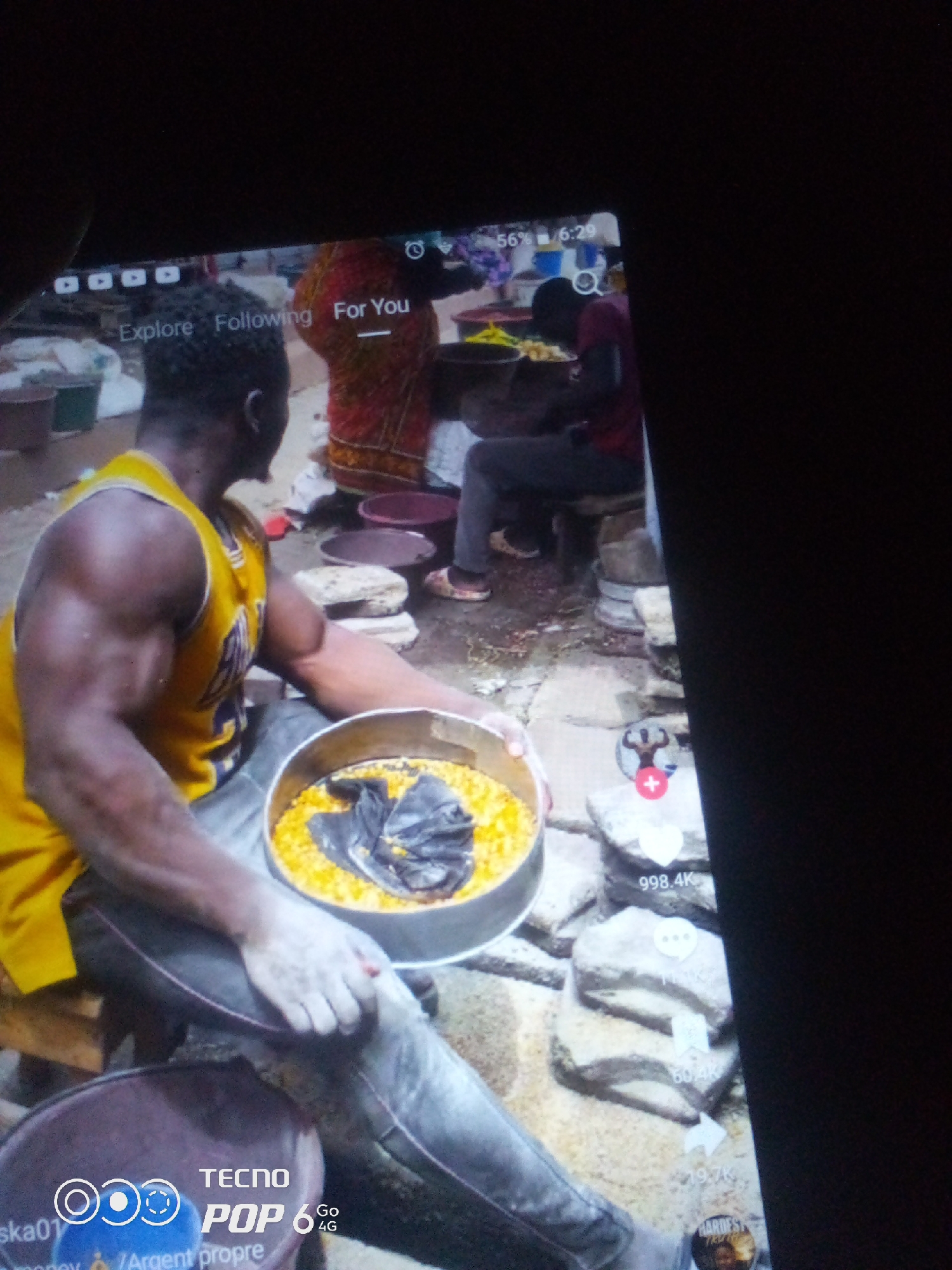 The image depicts a close-up view of a person holding a metal bowl filled with yellow corn kernels and a piece of black material. The person is wearing a yellow tank top and dark pants. In the background, other individuals are visible, along with various objects that suggest an outdoor market or food preparation area. The scene appears to be in a city, possibly Howayi, Nigeria, based on the text overlay in the lower left corner, which reads "TECNO POP 6 Go 4G." The overall setting is informal and likely involves a commercial activity related to food.
