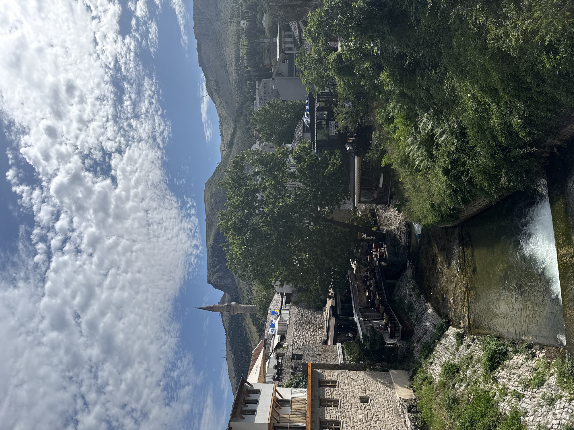 The image displays an outdoor scene in Mostar, Bosnia and Herzegovina, under a bright blue sky with scattered white clouds. A narrow river flows from the mid-ground towards the foreground, where a small weir generates white, aerated water. The riverbanks are lined with stone, partially covered by green vegetation.
On the left side of the river, a multi-story building constructed of light-colored stone features a balcony on its upper level. Behind and alongside this building, additional stone structures with visible windows and terraces are present. Several open-air restaurant or cafe areas, furnished with tables and chairs, are integrated into these riverside buildings, with a few individuals discernible as patrons. A prominent, slender, stone minaret with a pointed conical spire rises above the buildings on the left, slightly off-center. Below and to the left of the minaret, a blue flag with yellow triangles, identifiable as the flag of Bosnia and Herzegovina, is displayed on a building.
The right side of the image features dense green trees and foliage partially obscuring buildings and the riverbank. Further back, more buildings of varying architectural styles are visible.
The background is dominated by a range of green hills and mountains under a clear blue sky. Multiple white wind turbines are visible atop these distant elevations. The sky is characterized by dispersed, medium-sized white cumulus clouds. No specific actions or interactions are depicted beyond the passive presence of individuals in the restaurant areas.