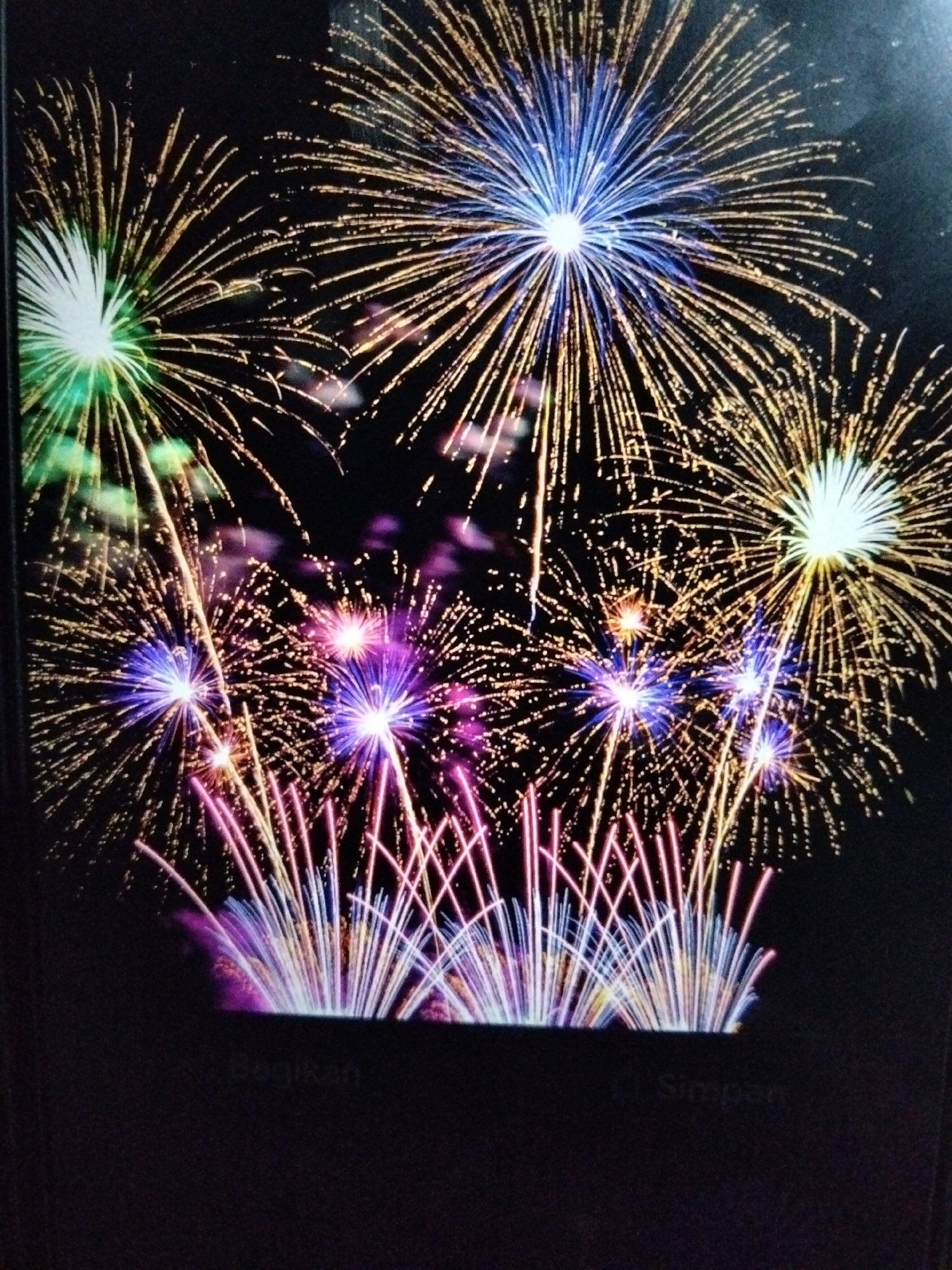 The image captures a vibrant fireworks display against a dark night sky, likely in Kualaraja, Indonesia, judging by the blurred text visible at the bottom of the frame, which appears to read "Regikan" and "Simpen." Multiple bursts of fireworks are visible, characterized by their radial patterns of light streaks. Colors include bright white, electric blue, vivid green, radiant pink, and golden yellow. Some fireworks have a dense, starburst effect, while others are more spread out, resembling weeping willows. The foreground is obscured by a dark area, possibly the edge of a screen or display, with faint reflections or text visible, further suggesting the image is from a digital source or a screen. No people or specific landmarks are discernible. The overall impression is one of celebration and spectacle.