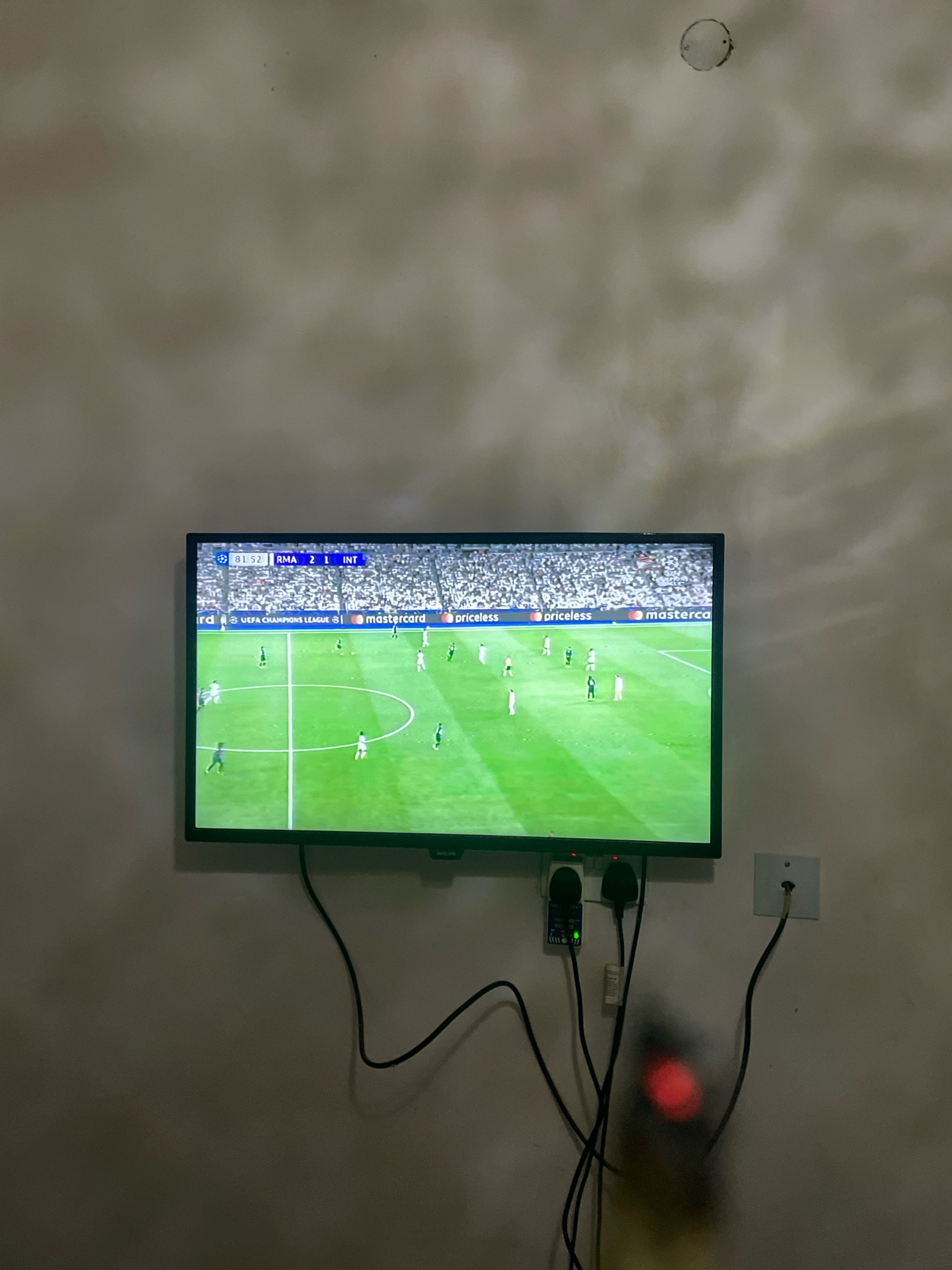 The image shows a flat-screen television mounted on a wall, displaying a soccer match. The score on the screen indicates "RMA 2 1 INT" at the 81st minute. Advertisements for UEFA Champions League, Mastercard, and Priceless are visible along the bottom of the screen. The wall appears to be painted in a light, neutral color and shows some texture and possibly shadows from lighting. Below the television, several electrical cords and adapters are plugged into wall outlets. A red light is visible on a device in the lower right portion of the image, partially obscured by the wires. The environment suggests an indoor residential or commercial space. Based on the image alone, the specific location in Abuja, Nigeria cannot be determined.