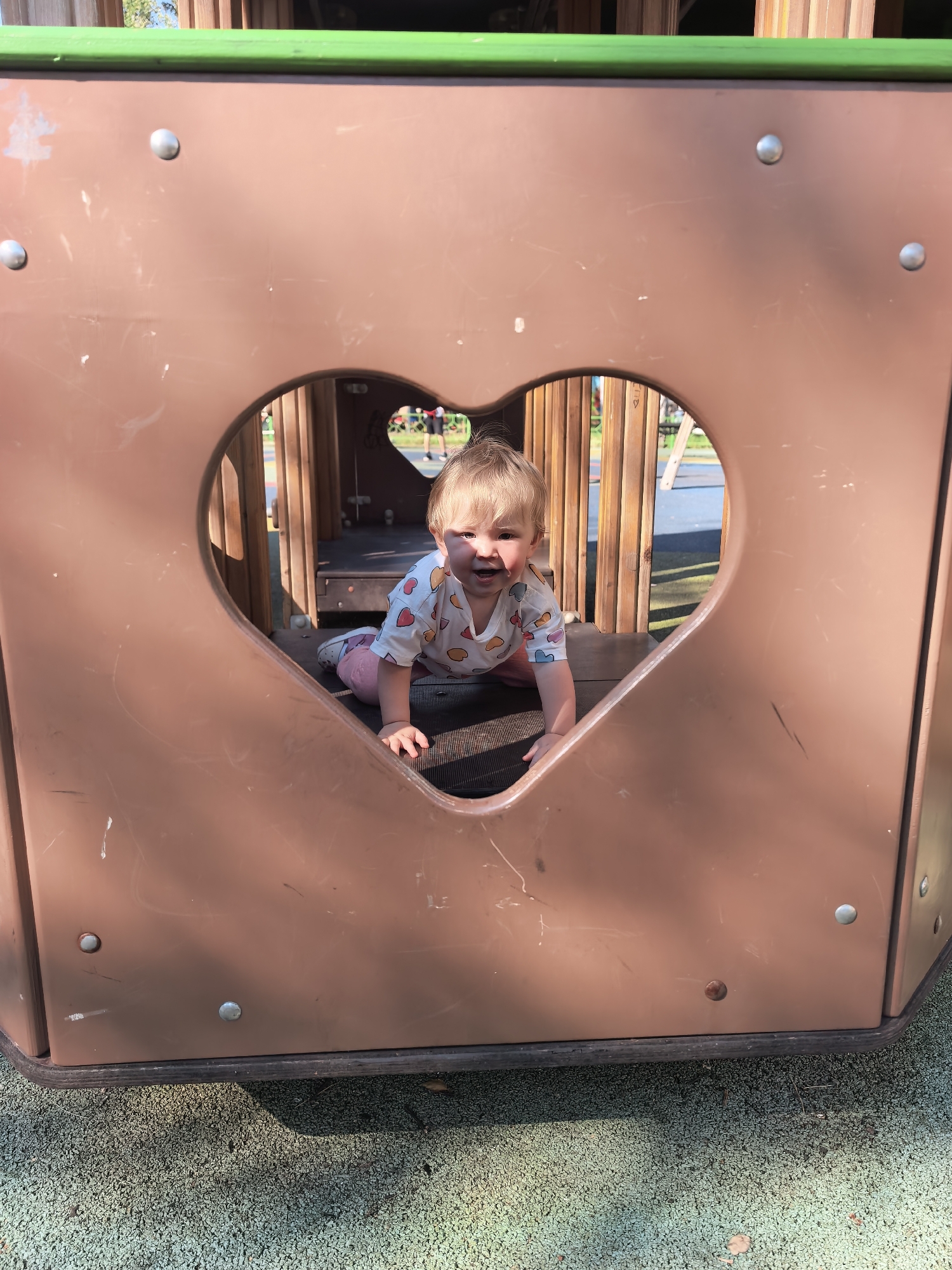 A young Caucasian child with blonde hair is crawling towards the camera through a heart-shaped opening in a brown plastic play structure. The child is wearing a white t-shirt with colorful heart patterns and pink pants. The play structure is outdoors, with wooden slats visible behind the heart opening and a green edge along the top. The ground appears to be a green rubberized surface, common in playgrounds. The image is taken from a low angle, looking up at the child. The scene suggests a sunny day, with shadows visible on the ground. The child is smiling and appears to be enjoying themselves. The image is a medium shot, with the child as the primary subject. The background is somewhat blurred, focusing attention on the child. The child's location is not specified in the image.
