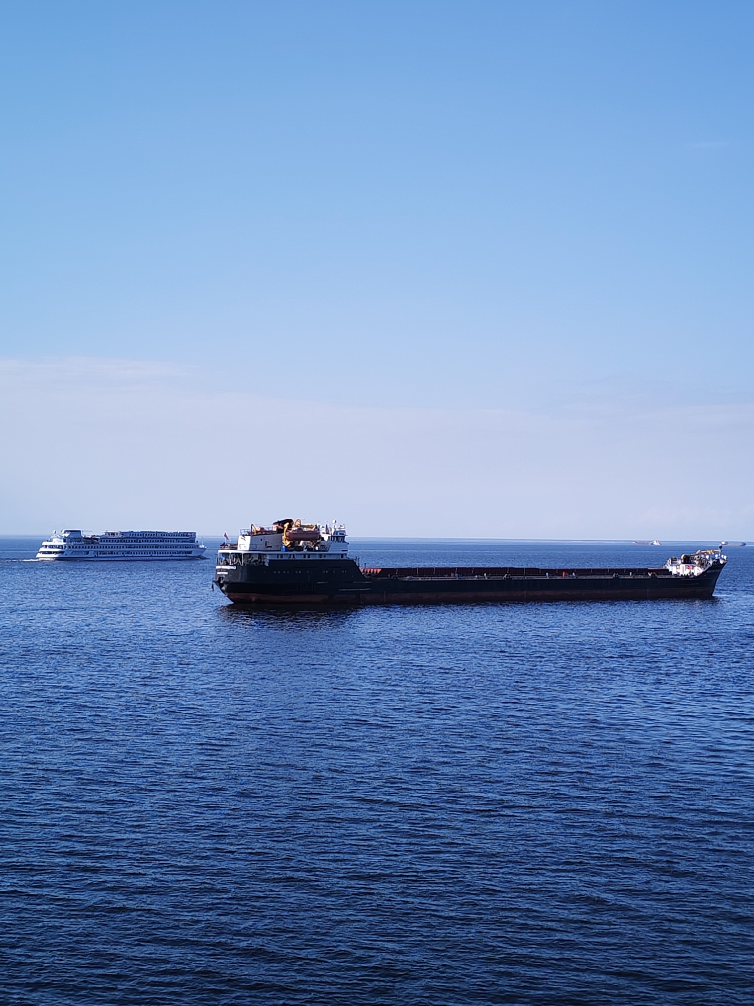 The image depicts a maritime scene. In the foreground, there is a long, dark cargo ship floating on blue water. It has a lighter-colored superstructure toward the front. To the left and further in the background, a white passenger cruise ship is visible. The sky above is predominantly blue with some faint clouds. The location is unknown, in the city of Unknown, Unknown.