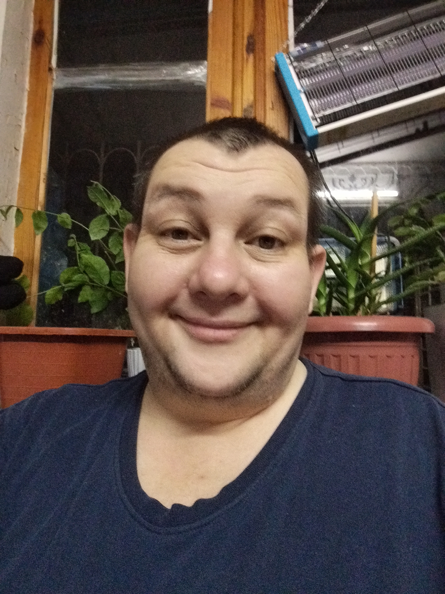 The image is a selfie of a light-skinned male with thinning brown hair and a beard. He is wearing a dark blue t-shirt. His expression is a wide smile, with his eyes slightly squinted. He is positioned in the foreground, with houseplants and what appears to be a window with wooden trim in the background. To the right of the man, there is an object that resembles an insect zapper hanging from the ceiling. The overall lighting is dim, suggesting an indoor setting at night. There is no identifiable information or context provided to place this image within the city of Tashkent, Uzbekistan.