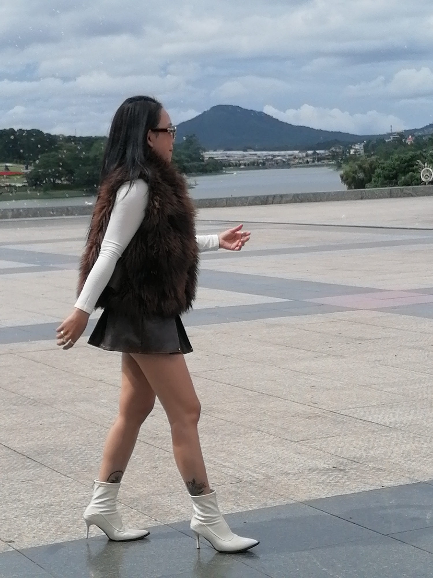 A woman with long dark hair and sunglasses is walking in a paved outdoor area with a body of water and a mountainous landscape in the background. She is wearing a white long-sleeved shirt, a dark fur vest, and a short dark skirt. She is also wearing white ankle boots. The area appears to be a public space, possibly a promenade or park, in Da Lat, Vietnam. The sky is cloudy.