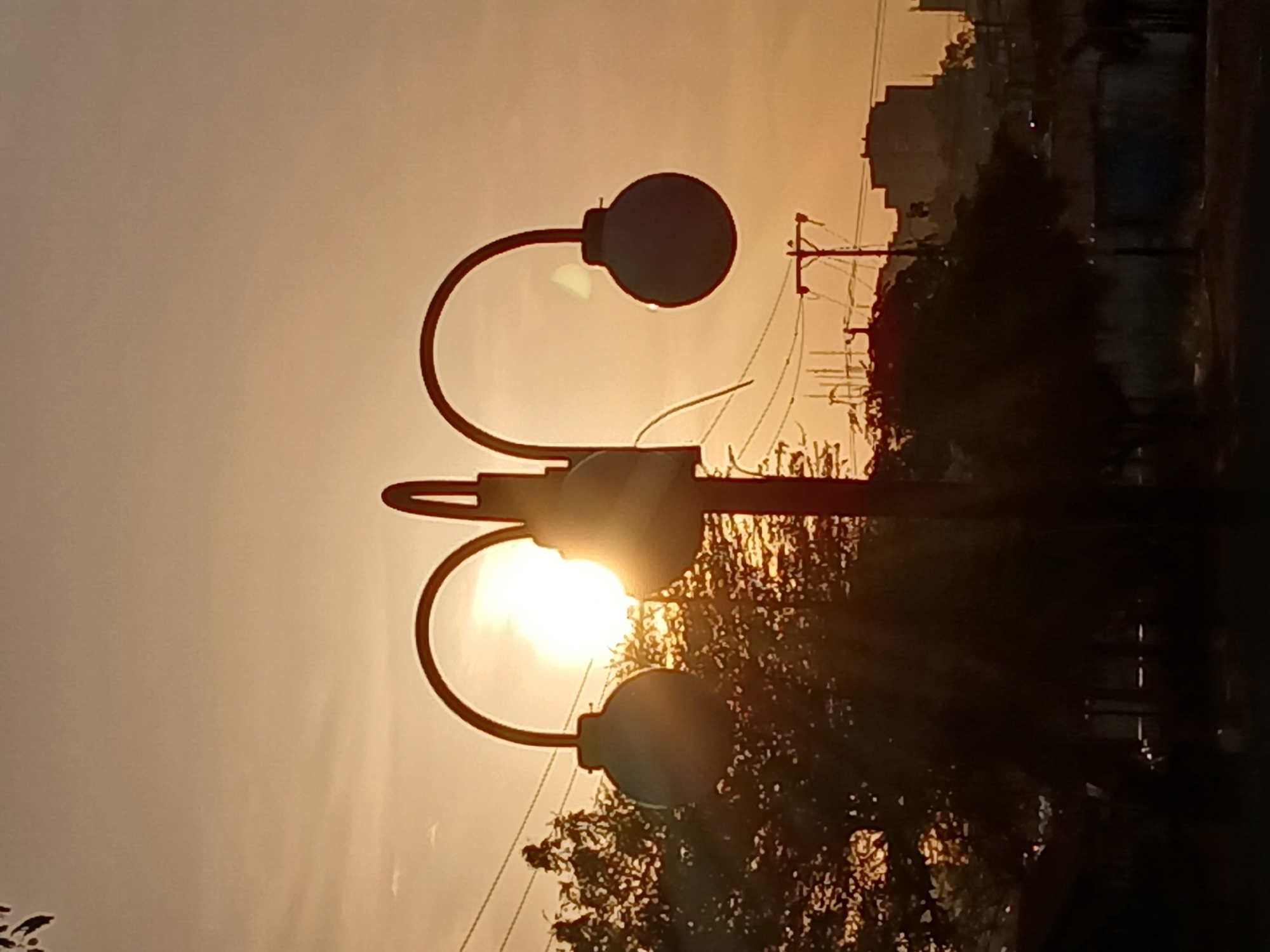 A street lamp with two spherical globes stands against a sunset sky in Ciudad Guayana, Venezuela. The sun is positioned behind the lower globe, creating a bright halo effect. Power lines are visible in the background, along with blurred silhouettes of buildings and trees. The image is slightly out of focus, with a warm, golden tone.