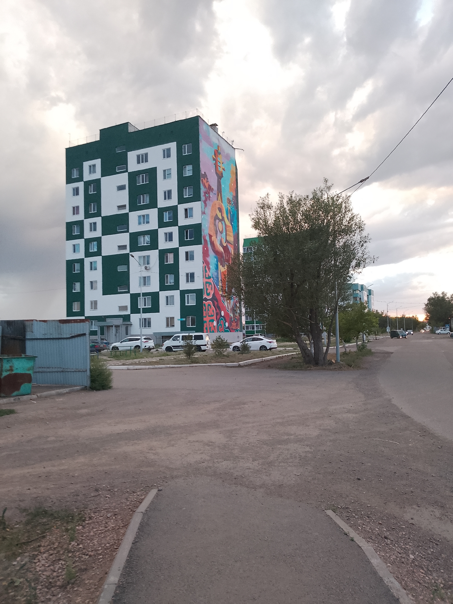 The image shows a street scene in the city of Abay, Kazakhstan. In the foreground, there is a dirt road with a curb. To the left, there is a metal fence with a trash bin. In the middle ground, there is a tall, modern apartment building with a green and white checkered facade and a colorful mural on its side. Several cars are parked in front of the building. A large tree stands in front of the building, partially obscuring it. In the background, there are more apartment buildings, streetlights, and a power line stretching across the sky. The sky is cloudy and overcast, with a hint of sunset colors peeking through. The overall scene depicts a residential area in a city under a twilight sky.