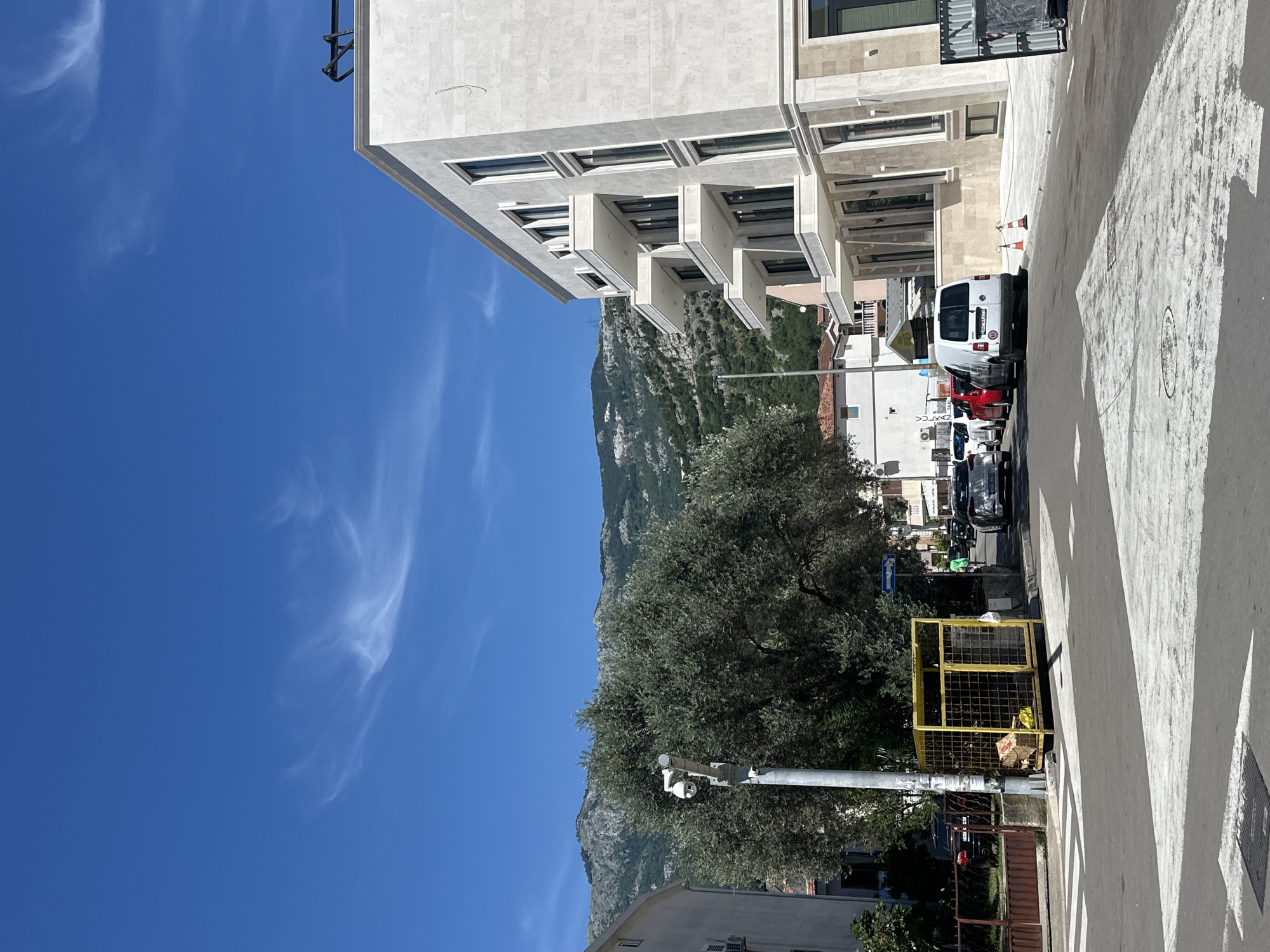 This is a street-level photograph taken in Budva, Montenegro, on a bright, sunny day. The scene depicts a residential street with modern buildings, vehicles, and natural elements.
In the foreground, a white arrow painted on the asphalt indicates a turn or lane direction. To the left, a yellow metal cage filled with debris stands on the sidewalk, next to a light pole with a security camera attached. A large, mature olive tree with dense foliage dominates the left-middle ground.
The middle ground features several parked and moving vehicles. A white van is parked on the right side of the street, with a red car and other vehicles visible behind it. A person in a green shirt is walking on the sidewalk further down the street.
The background is characterized by a range of buildings, including a modern, multi-story building on the right with balconies and large windows. Behind the buildings and trees, rolling green hills and mountains rise against a clear, bright blue sky with wispy white clouds.
The overall impression is one of a well-maintained, urban environment with a Mediterranean flair, likely a residential or commercial area within the city of Budva.