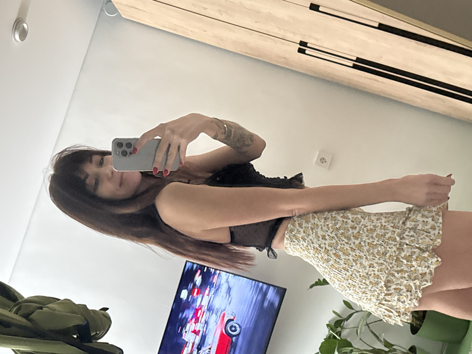 The image displays a mirror selfie taken by a woman, rotated 90 degrees counter-clockwise so that the woman appears to be lying on her back, facing upwards.
The woman is positioned in the center, holding a light grey smartphone with multiple rear cameras in front of her face with both hands, using it to capture her reflection in the mirror. She has long, dark brown hair with bangs, falling down around her shoulders. Her fingernails are painted red. A tattoo is visible on her right upper arm (left side of the image from her perspective). She is wearing a black, fitted top, possibly a bustier or corset style, with a slightly sheer or textured material, and a light-colored (off-white or cream) mini-skirt with a small yellow floral or patterned print. The skirt appears to have shirring or ruching texture. Her expression is neutral with a slight hint of a smile.
The background, which is the reflection of the room, shows:
* A plain white wall filling most of the upper and lower portions of the image.
* In the upper right (which would be the top right of a correctly oriented image), a wooden dresser or cabinet with two prominent black horizontal handles is visible.
* In the lower left (which would be the bottom left of a correctly oriented image), a flat-screen television is displaying an image of what appears to be a racing game or video, featuring a red car.
* Next to the television, in the very bottom left corner, part of an olive green or khaki-colored bag or backpack is visible.
* In the lower right (bottom right of a correctly oriented image), a green potted plant with large leaves is partially visible.
* A white electrical outlet is visible on the white wall between the woman and the wooden furniture.
The lighting in the room appears bright and even, suggesting either daytime or well-lit indoor conditions. There is no visible text in the image.