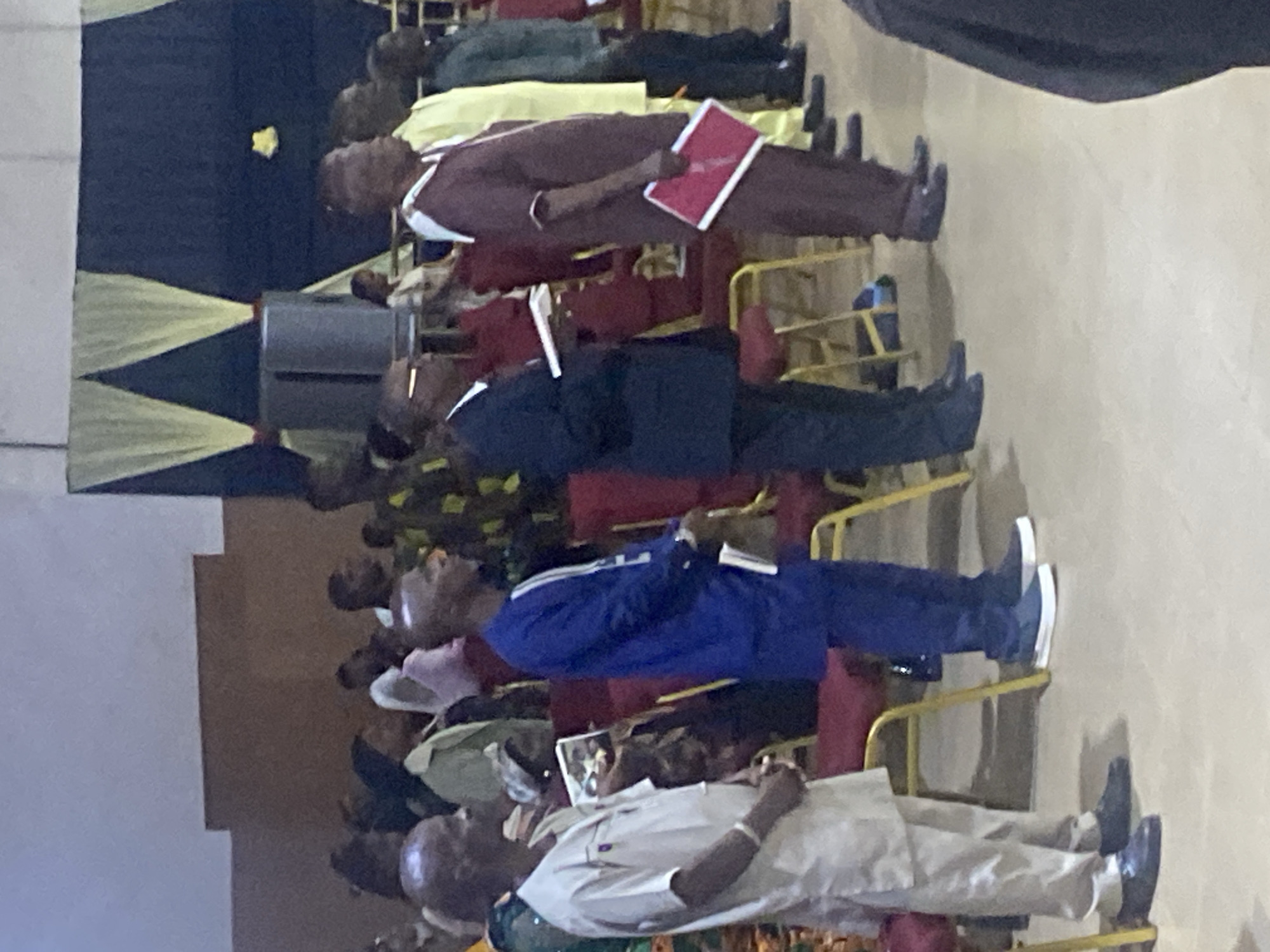 A group of men are standing in a line indoors, facing to the right of the frame. The man in the foreground on the left is wearing a light-colored robe and pants. Next to him, a man in a blue robe and pants holds a white booklet. To his right, a man in a dark suit holds a white booklet. Further down the line, a man in a maroon robe and pants holds a red folder. Behind him, a man in a yellow robe and pants stands, followed by a man in a dark green robe and pants. The men are standing on a light-colored floor in front of red chairs. A dark blue curtain with yellow drapes hangs in the background. A gray speaker is visible to the left of the curtain. The scene appears to be at an event or service. The location is likely Rukuba Road, Nigeria, based on the provided context.