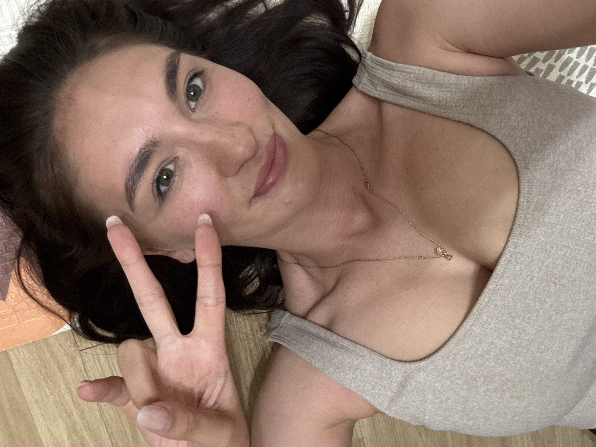 A young woman is lying down, taking a selfie at an angle. She has dark hair, fair skin, and is wearing a light-colored tank top and a gold necklace. Her left hand is raised in a "peace" sign. The background appears to be a floor with a rug or mat partially visible. The image does not provide sufficient information to determine if it was taken in Sochi, Russia, or any other specific location.