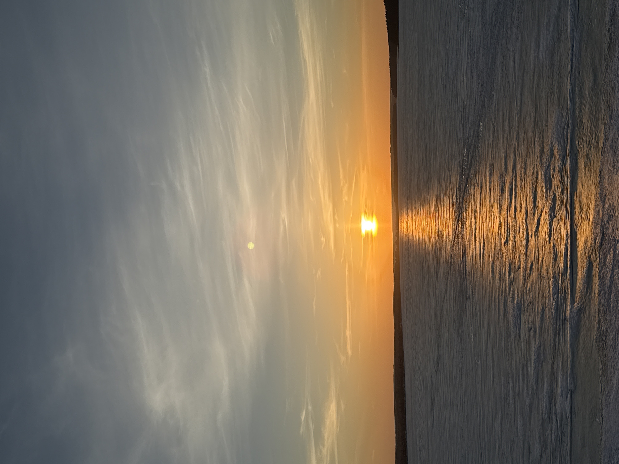 The image shows a sunset over a body of water, possibly a lake or a river. The sun is low on the horizon, casting a bright orange glow across the sky and reflecting on the water's surface. There are clouds in the sky. The body of water is partially obscured by some vertical linear textures on the right side. There is no information to confirm if the image was taken at Votkinsk, Russia.
