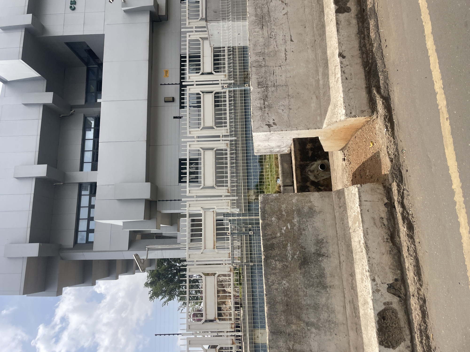 The image depicts an urban scene in Jos, Nigeria. A modern, multi-story building with a boxy, geometric design dominates the left side of the frame. The building is primarily white or light gray in color and features multiple windows with dark glass.
A white metal fence runs parallel to the building. A concrete barrier or wall is visible between the fence and the road. The concrete is weathered, with visible cracks and stains. A drainage ditch is present along the road, with a pipe visible within it. The road surface is asphalt with a yellow dividing line. The sky is visible in the lower left, with white clouds.