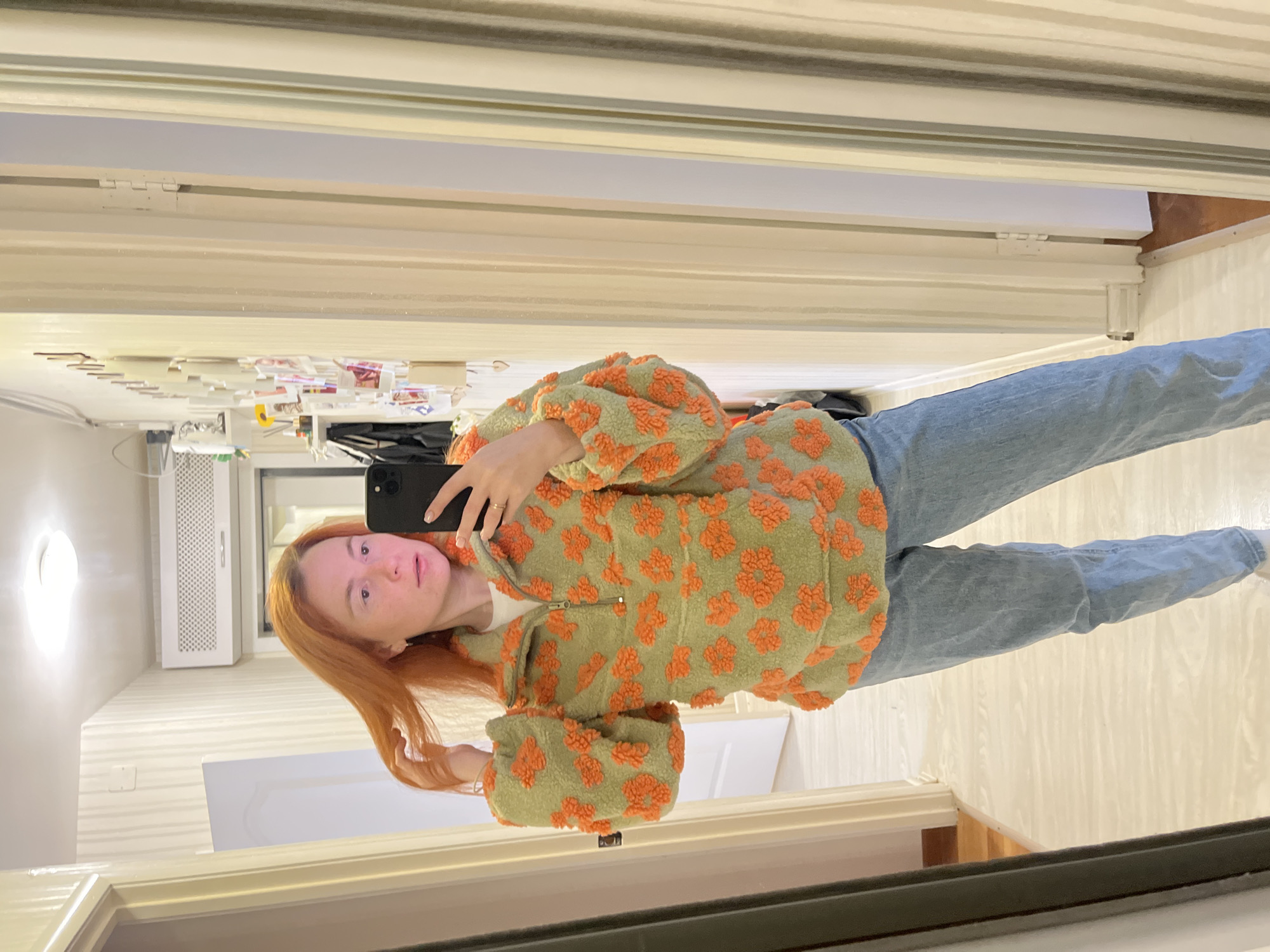 A young woman with auburn hair is taking a mirror selfie in what appears to be a hallway. She is wearing a light green fleece pullover with an orange floral pattern and light blue jeans. The hallway has pale wood floors and white paneled walls. To the right of the woman, a tall mirror reflects her and a section of the hallway. On the wall in the reflection, there are several framed photos and some decorative items. The lighting in the hallway is bright, likely from overhead fixtures. The overall scene suggests a casual, domestic setting. No specific location in Chaikovsky, Russia, can be determined from the visual information provided.