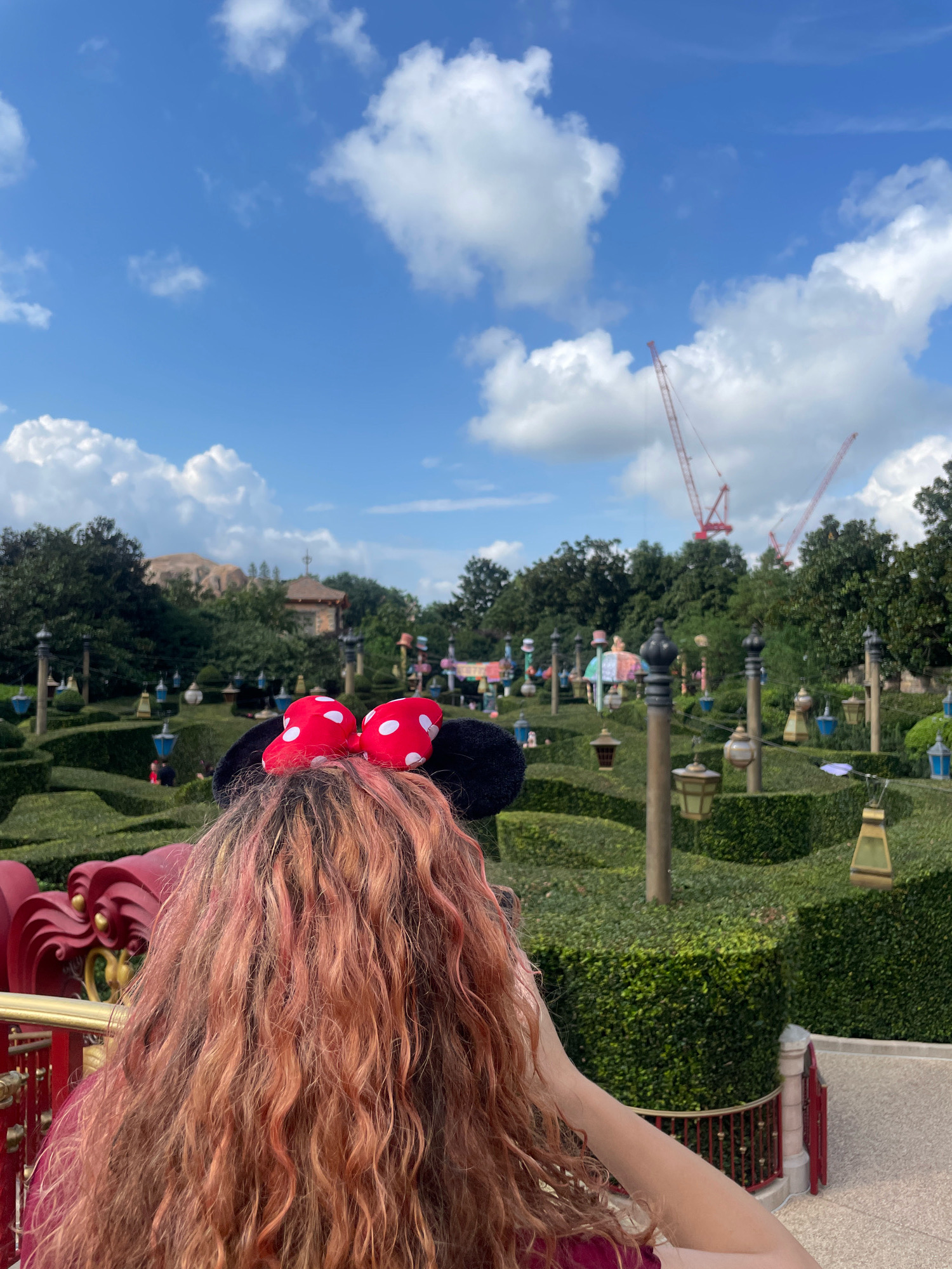 In Pudong, China, a person with long, wavy, pink-tinged hair, wearing Minnie Mouse ears with red and white polka dot bows, is viewed from behind. They are leaning on a red railing with gold accents, overlooking a meticulously manicured hedge maze. The maze features numerous lampposts with lanterns in various colors, including blue, yellow, and white, suspended by wires. In the background, lush green trees form a dense canopy, with a building visible behind them. The sky above is a bright blue, dotted with fluffy white clouds. Two large red construction cranes are prominently visible in the distance, suggesting ongoing development. The scene captures a moment at an amusement park or a similar recreational area.