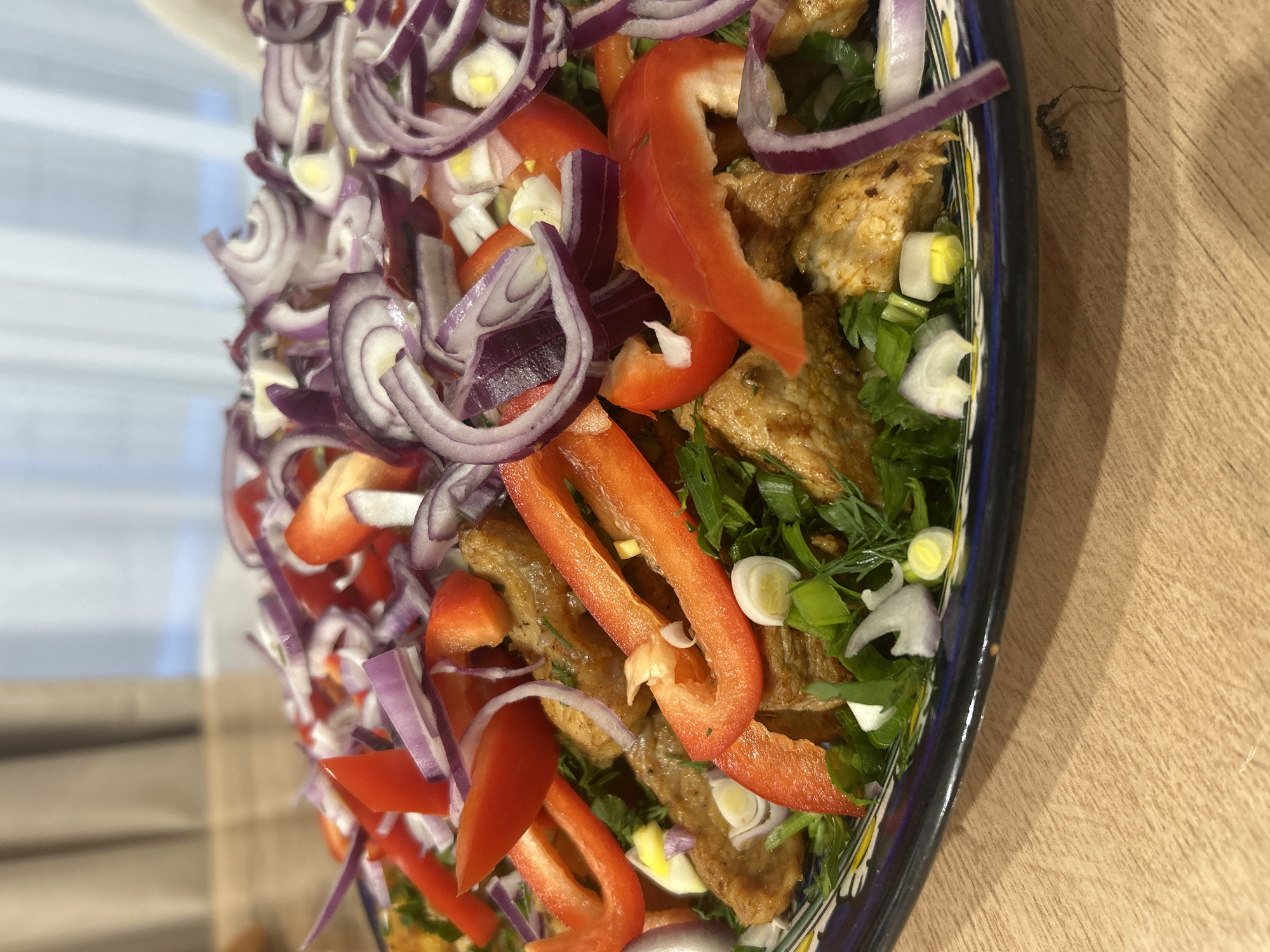 A close-up, angled shot of a plate of salad is presented on a wooden surface. The salad comprises sliced red onions, strips of red bell pepper, pieces of cooked chicken or meat substitute, chopped green onions, herbs (possibly dill and parsley), and small white pieces which may be cheese or hard-boiled egg. The plate itself has a blue and yellow decorative border. The background is partially obscured, showing a window with a blurry, indistinct view. The overall setting suggests a meal preparation scene, likely indoors. There is no evidence to indicate this image was taken in Chaikovsky, Russia.