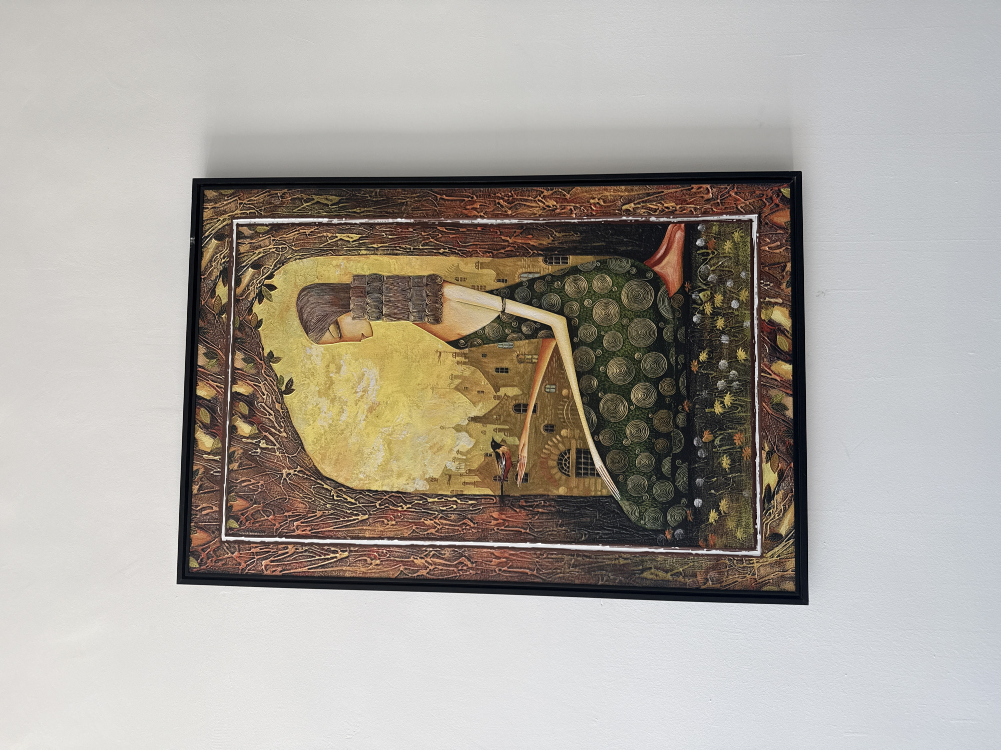 A framed painting hangs on a white wall. The painting depicts a woman with a stylized, elongated figure lying horizontally within a landscape. She has brown hair and wears a headdress or collar composed of patterned squares. Her skin is a light yellow color. Her dress is long and green, decorated with gold spirals. In the background of the painting, behind the woman, there's a cityscape with stylized buildings in yellow and brown tones. A small red figure stands near the center of the landscape. The painting is framed with a brown, textured border that resembles branches and leaves. The frame is dark and hangs centered on the white wall. There is no evidence of Izhevsk, Russia in the image.