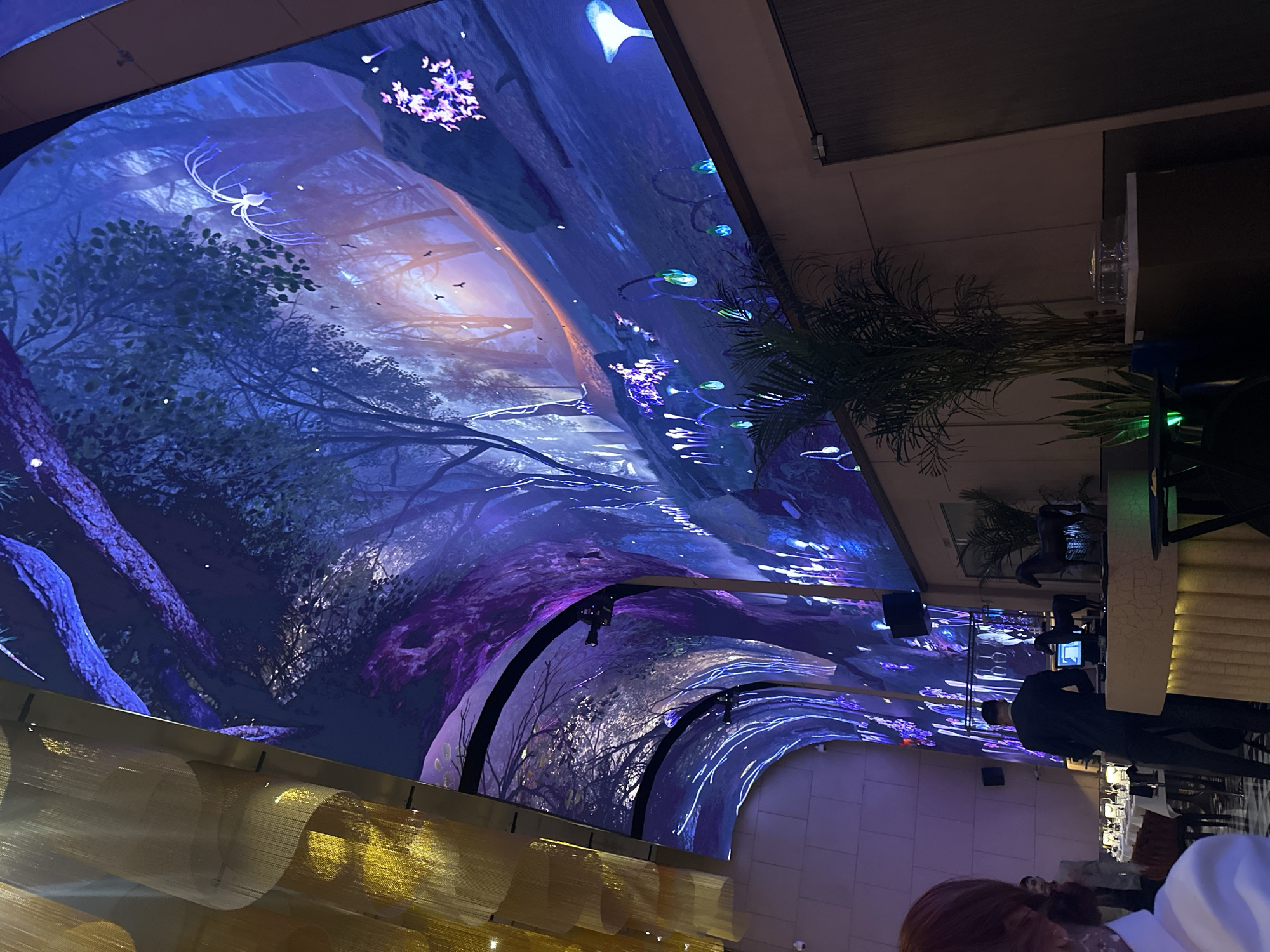 This image captures an opulent and immersive indoor setting, likely a high-end restaurant or event venue in Dubai, United Arab Emirates. The dominant feature is a massive, curved digital display that covers the ceiling and upper walls, creating an enveloping, tunnel-like environment.
The digital display projects a vibrant, fantastical forest scene, highly reminiscent of the bioluminescent world depicted in "Avatar." The scene is rich with detail, featuring intricate tree branches, glowing flora in shades of blue, purple, and green, and small, ethereal flying creatures. Notable elements within the projection include white, jellyfish-like organisms with long tendrils, clusters of purple-pink glowing "flowers," and various other bioluminescent plants that illuminate the digital landscape.
In the foreground on the left, shimmering golden-toned decorative panels or light fixtures add a touch of luxury to the space. On the right side of the image, the real-world environment becomes visible. A large, leafy palm plant stands near the edge of the digital display. Further down, several individuals are present, suggesting a social gathering or dining experience. One person in a dark suit is standing, partially obscured by a structural element, while another person's head with reddish-brown hair is visible in the bottom right corner.
The lighting in the physical space is dim, allowing the vivid digital projection to be the primary source of illumination and the focal point, indicating an evening or night setting. The overall atmosphere is one of wonder and futuristic enchantment. There is no visible text in the image.