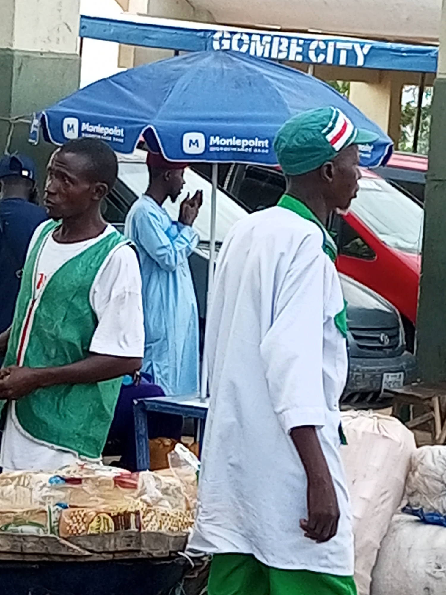 This image depicts a street scene in Gombe City, Nigeria. In the foreground, a man wearing a green vest over a white t-shirt stands facing left, looking away from the camera. To his right, another man in a white robe and a green, white, and red striped cap is also facing left. Behind them, a man in a light blue robe stands with his hands clasped, looking down. A blue umbrella with "Moniepoint" logos is open above them. In the background, vehicles, including a red car and a dark-colored SUV, are visible. The environment suggests an outdoor market or street-side vendor area, with bags of goods and a table displaying items. The overall lighting indicates it is daytime.