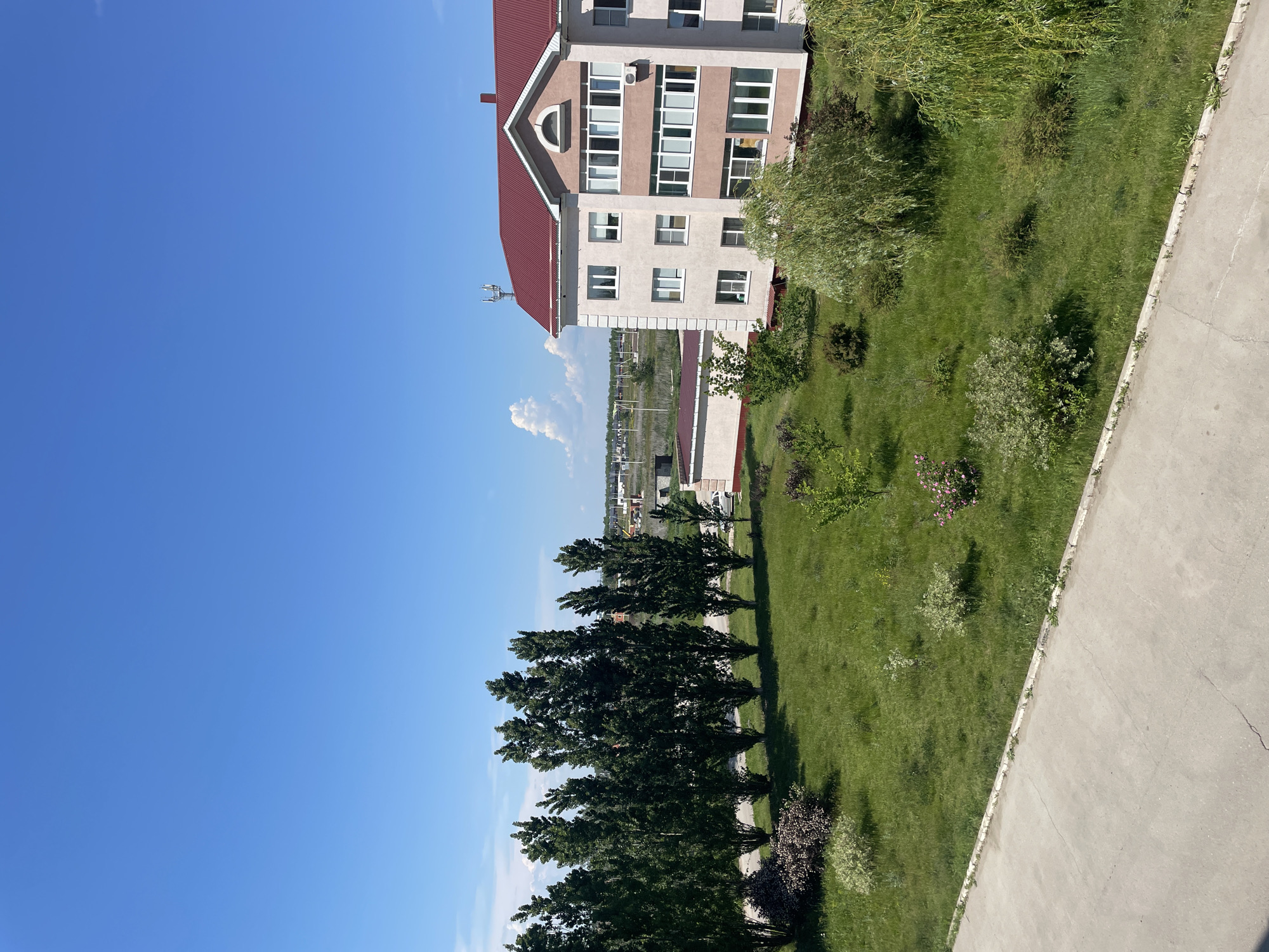 A medium shot captures a clear, bright blue sky with a few wispy clouds on a sunny day. In the foreground, a concrete path stretches diagonally from the bottom left to the bottom right of the frame. Beyond the path is a lush green lawn dotted with various trees and bushes, some of which are in bloom with pink flowers. A row of tall, slender trees stands prominently on the left side of the lawn, casting shadows. On the right side of the image, a multi-story building with a red roof is visible. The building has beige and pink facades, with numerous windows and balconies. A cellular tower is mounted on the roof. In the background, more buildings and structures are sparsely scattered across the landscape. The scene appears to be an outdoor area in a residential or urban setting.
