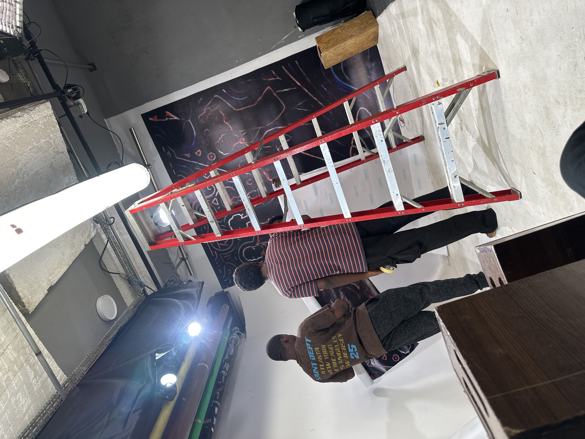 An indoor scene, likely a studio or workshop in Ikeja, Nigeria, captures two men engaged in setting up or working with equipment. The image is oriented vertically but depicts the subjects standing on a floor.
In the foreground, a large red and silver stepladder is prominently positioned. Behind it, a black banner featuring abstract red and white scribble-like patterns is affixed to a wall. Two men, both appearing to be Black, are visible. One man wears a red and white striped short-sleeved shirt and black pants, standing near the ladder. The second man, positioned further right, wears a brown long-sleeved shirt and dark grey sweatpants.
The setting is characterized by studio lighting equipment, including a long, bright white light on the left and other bright spotlights, indicating an active work environment. On the left side of the frame, there's a light gray textured wall or backdrop. To the right, dark brown wooden boxes or shelves are visible. A roll of white material, possibly a backdrop, is also present, with the text "ABRO" discernibly printed on its packaging. Other items include wires, cables, and a black bag.
The brown shirt worn by one of the men displays the text: "SAINT DEPT", followed by a list of cities "ATLANTA", "NEW YORK", "CHICAGO", "LOS ANGELES", "NEW JERSEY", and the number "25". Additional text partially visible on other objects, as per OCR, includes "ABRD", "BRO", "DHORT", "CHAPU", and "OMER". The overall atmosphere suggests a functional, utilitarian space where the men are likely involved in a setup or installation task. The time of day is indeterminate due to artificial lighting.