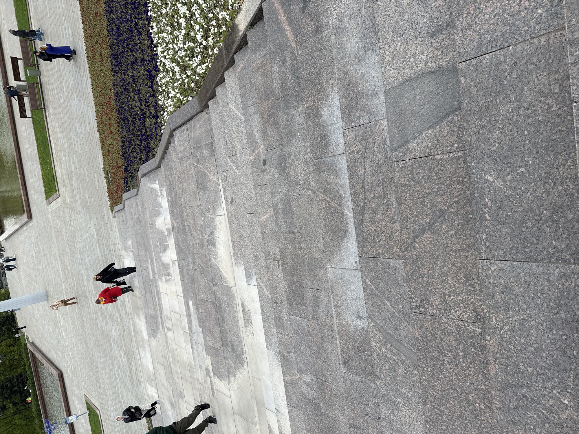 The image shows an elevated, angled view of a public space, likely in a city, with many pedestrians.
**Location:** The city is identified as Moscow, Russia.
**Environment:** The ground is paved with light gray tiles, possibly granite or similar material. There is a staircase of the same material on the right side of the image. Next to the staircase there is an area of vegetation, including flowers. There is also a water feature.
**People:** Multiple individuals are visible. They are dressed in various styles of clothing and appear to be walking around the area. Some are wearing coats or jackets, suggesting a mild or cool temperature.
**Objects:** There are benches alongside the water feature and other urban elements such as street signs.