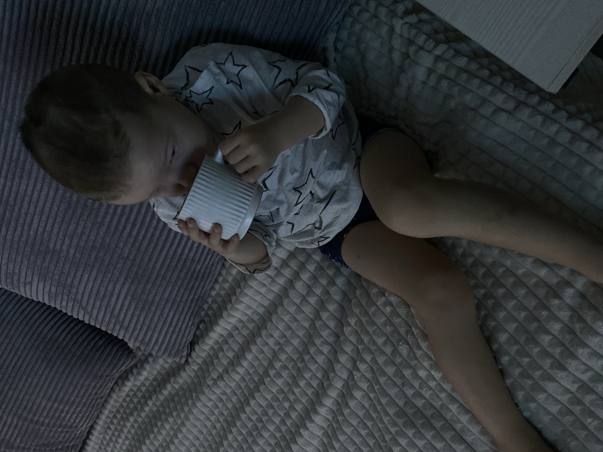 This image depicts a young child seated on a bed in a dimly lit indoor environment, identified as being located in Sochi, Russia. The child, with light brown hair, faces slightly left and is actively drinking from a white, vertically ribbed cylindrical mug, held with both hands. The child wears a light-colored, long-sleeved shirt patterned with dark stars and dark shorts or underwear. The bed is covered by a light gray, textured blanket with a wavy pattern. Two dark gray, horizontally ribbed pillows are positioned against what appears to be a headboard or wall behind the child. A light-colored, flat surface, possibly a bedside table or shelf, is partially visible in the lower right corner. The overall lighting suggests an evening or nighttime setting.