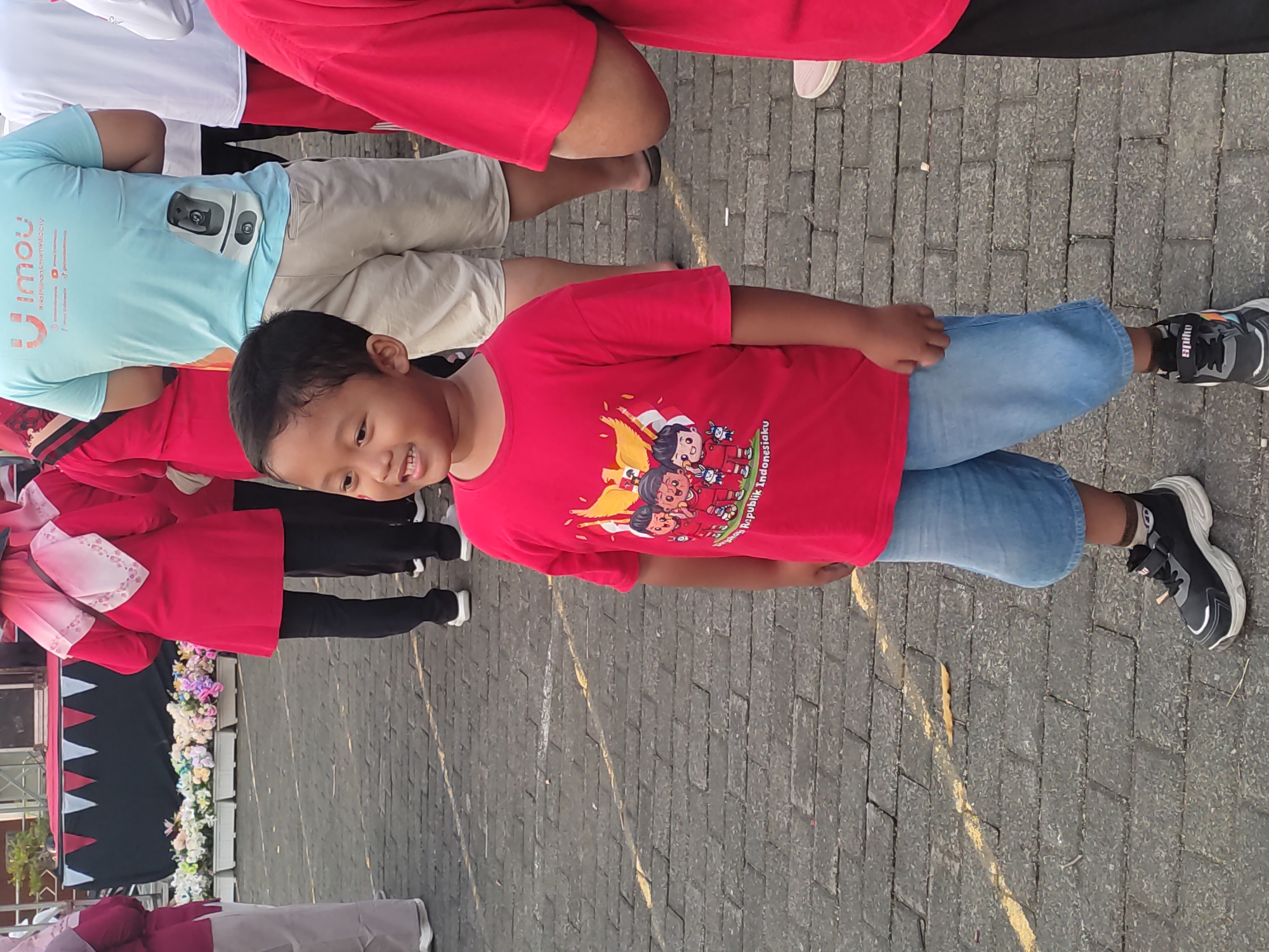 A young Indonesian boy, standing on a cobblestone pavement, is the central subject of this outdoor photograph. He wears a red t-shirt with a graphic depicting cartoon characters and text that appears to say "Republik Indonesiaku," and light blue denim shorts. His feet are clad in black and white sneakers. The boy is looking to his left with a slight smile.
In the background, several other individuals are visible, many wearing red attire. To the boy's left, a group of women in matching red outfits and headscarves stand in front of a stage decorated with a black and red banner and a row of white planters filled with colorful flowers. To the boy's right, an adult wearing a red shirt and black pants is partially visible. Further in the background, an individual in a light blue t-shirt with a logo and text is also present.
The setting appears to be an outdoor public space, possibly a festival or event, given the stage and the gathered crowd. The cobblestone ground is marked with yellow lines. The overall atmosphere suggests a daytime event in a populated urban or semi-urban area within Ponorogo, Indonesia.
