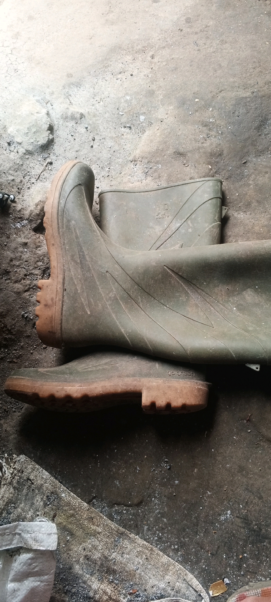 Two olive green rubber boots are placed on a rough, concrete floor. The boots are oriented diagonally in the frame, with the toe of the lower boot pointing towards the bottom left corner, and the top of the upper boot extending towards the top right. The lower boot is resting on its side, with its sole clearly visible. The sole has a thick, lugged tread pattern. The upper boot is positioned on top of the lower boot, its shaft extending upwards. The material of the boots appears worn and dusty. There are visible embossed logos on the boots; one appears to say "WP BOOTS" and another "GO GREEN". The concrete floor is dirty and uneven, with scattered debris and some larger stones. In the bottom left corner, a piece of white woven material, possibly a bag, is visible next to a textured wooden surface. The lighting suggests an indoor setting, possibly a workshop or storage area, with shadows cast on the floor. The location is specified as Penanggungan, Indonesia, though there are no specific visual cues in the image to confirm this location.