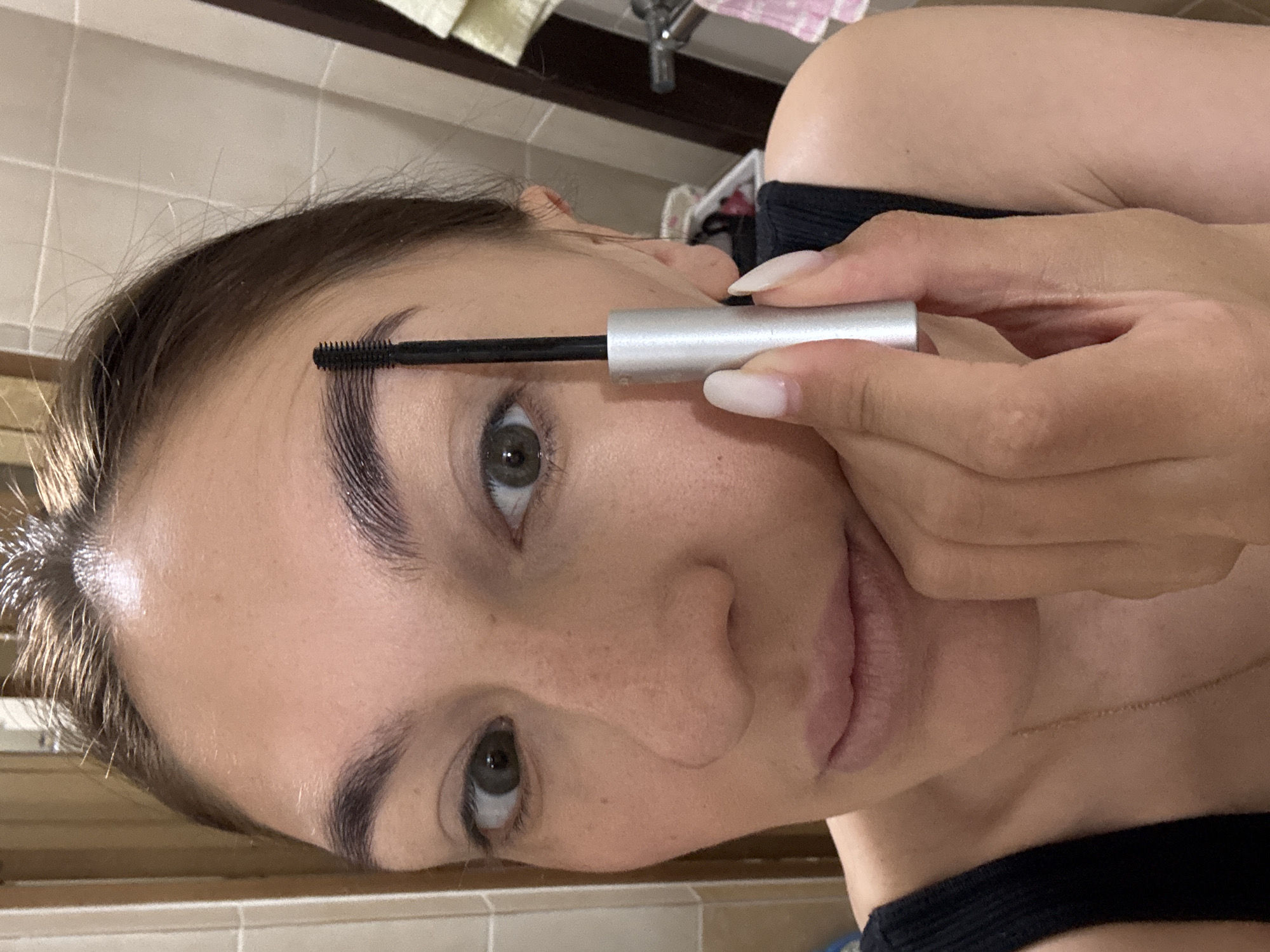 A woman applies eyebrow gel in a bathroom. She is holding a silver tube and brush to her left eyebrow. The background includes a tiled wall, a towel, and a sink. There is no information to indicate this image was taken in Sochi, Russia.