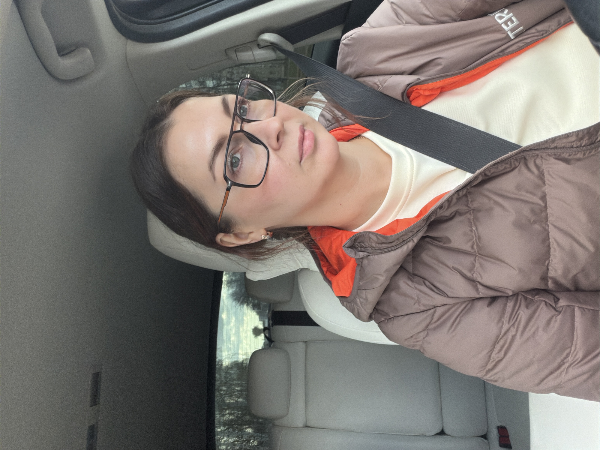 An eye-level portrait shows a young woman sitting in the passenger seat of a car, looking directly at the camera. She has medium-brown hair, glasses with black frames and orange/brown temples, and a neutral expression. She wears a light cream or off-white long-sleeved shirt under a brown puffer jacket with a bright orange lining. A black seatbelt is diagonally across her chest and shoulder.
The car's interior is visible, featuring a grey headliner and light-colored (possibly white or light grey) leather seats. A grey grab handle is on the ceiling above her. Through the window, outside the car, blurred trees and what appears to be an overcast sky suggest daytime and cooler weather. A partial view of "TER" is visible on the collar area of her jacket, indicating the brand "ARC'TERYX". The location context provided is Chaikovsky, Russia.