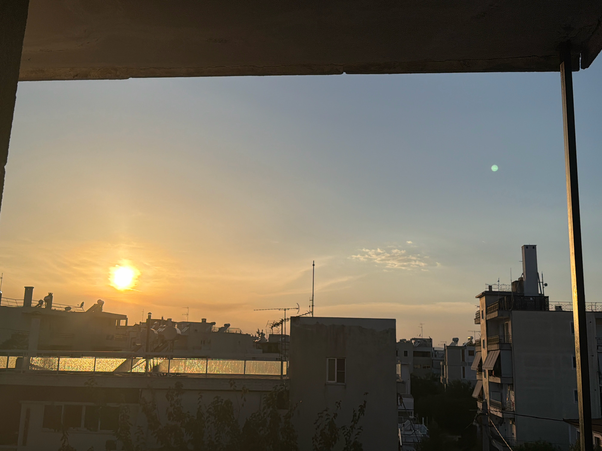 The image captures a sunset over the city of Χαλάνδρι, Greece, viewed from an elevated position, likely a balcony. The sun, a bright orb casting a warm, golden light, is low on the horizon to the left, illuminating the sky with hues of orange and yellow. wispy clouds are scattered across the sky. In the foreground, the dark silhouette of a concrete structure, possibly a balcony or overhang, frames the upper part of the scene. Below this, rooftops of various apartment buildings are visible, characterized by their white or light-colored facades. Many buildings feature balconies, awnings, and rooftop antennae. A distant, faint green circular object is visible in the sky to the right. The overall impression is an urban landscape bathed in the soft, fading light of sunset.