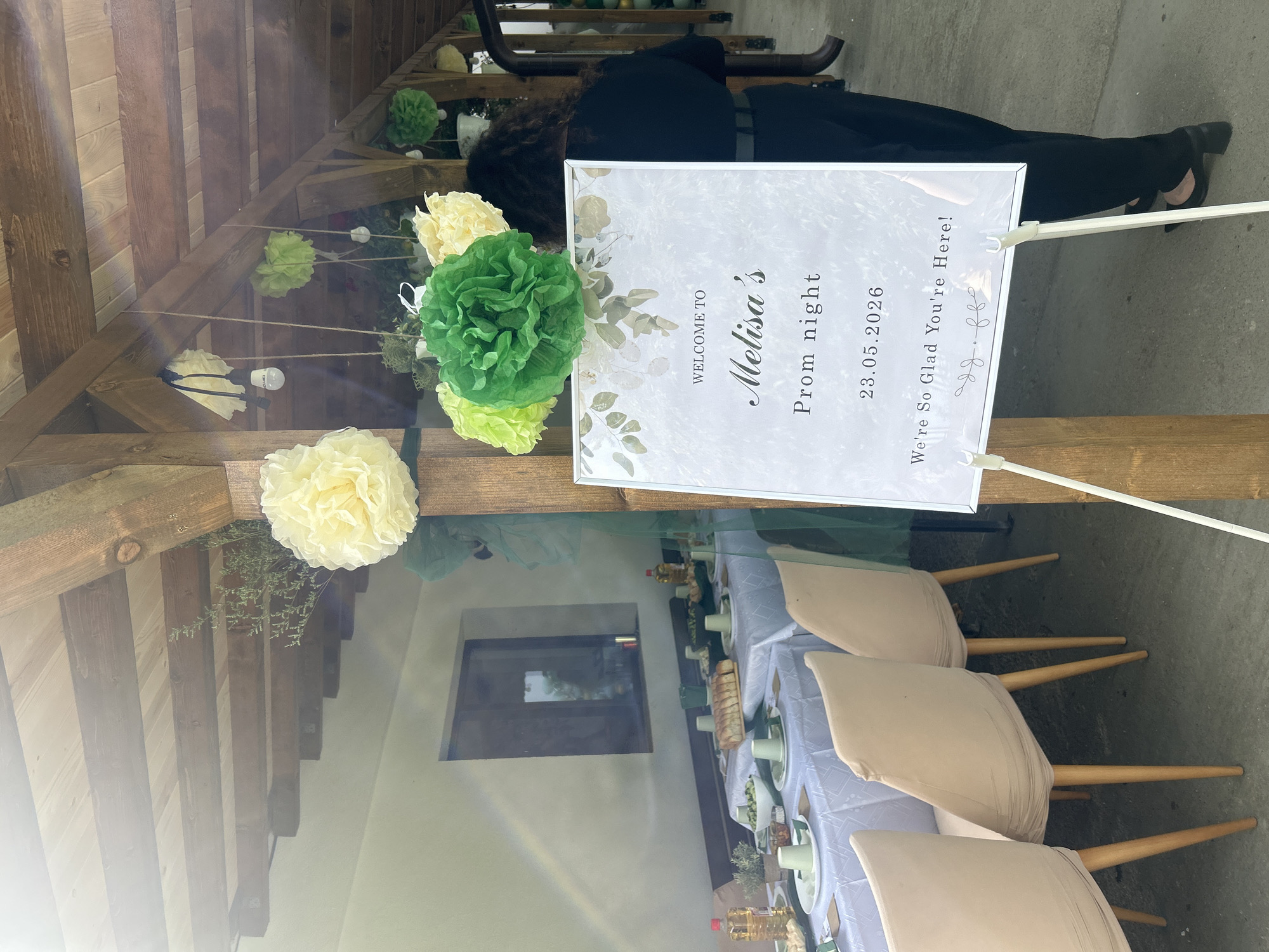 The image depicts an outdoor event, possibly a prom night celebration, in what appears to be a covered patio or open-air structure. The wooden structure is decorated with paper flowers and greenery. A sign reads "Welcome to Melisa's Prom night 23.05.2026 We're So Glad You're Here!"
A person in black clothing is visible behind the sign.
A table setting with beige chairs, a white tablecloth, and tableware is visible. The location is identified as Sandrovo, Bulgaria.