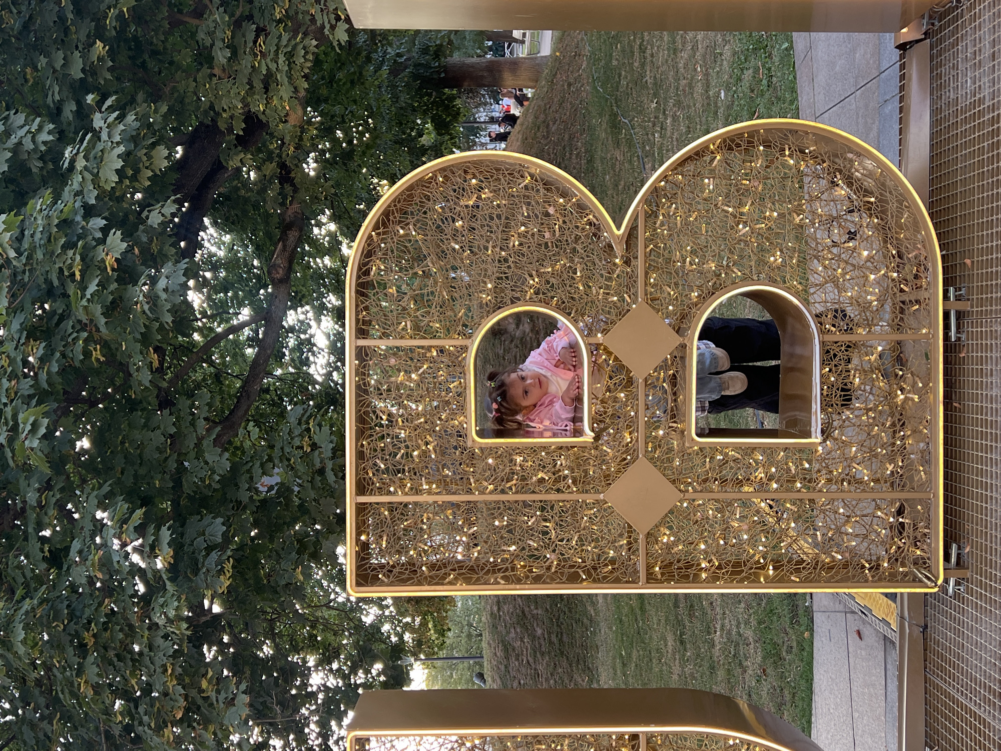 The image shows a large, gold, illuminated decorative letter "B" in the city of Lublin, Poland. The letter is filled with a tangled network of warm-toned string lights. A young child with pigtails, wearing a pink jacket, is peeking out of an opening in the letter. Below the child, a person's legs and feet wearing blue jeans and brown shoes are visible through another opening. The letter is positioned outdoors on a grid-like surface, with a grassy area and trees in the background. Sunlight filters through the leaves of the trees. In the distance, other people are visible, some sitting on a grassy hill and others on benches. The overall scene appears to be a public space, possibly a park or plaza, decorated for an event or celebration.