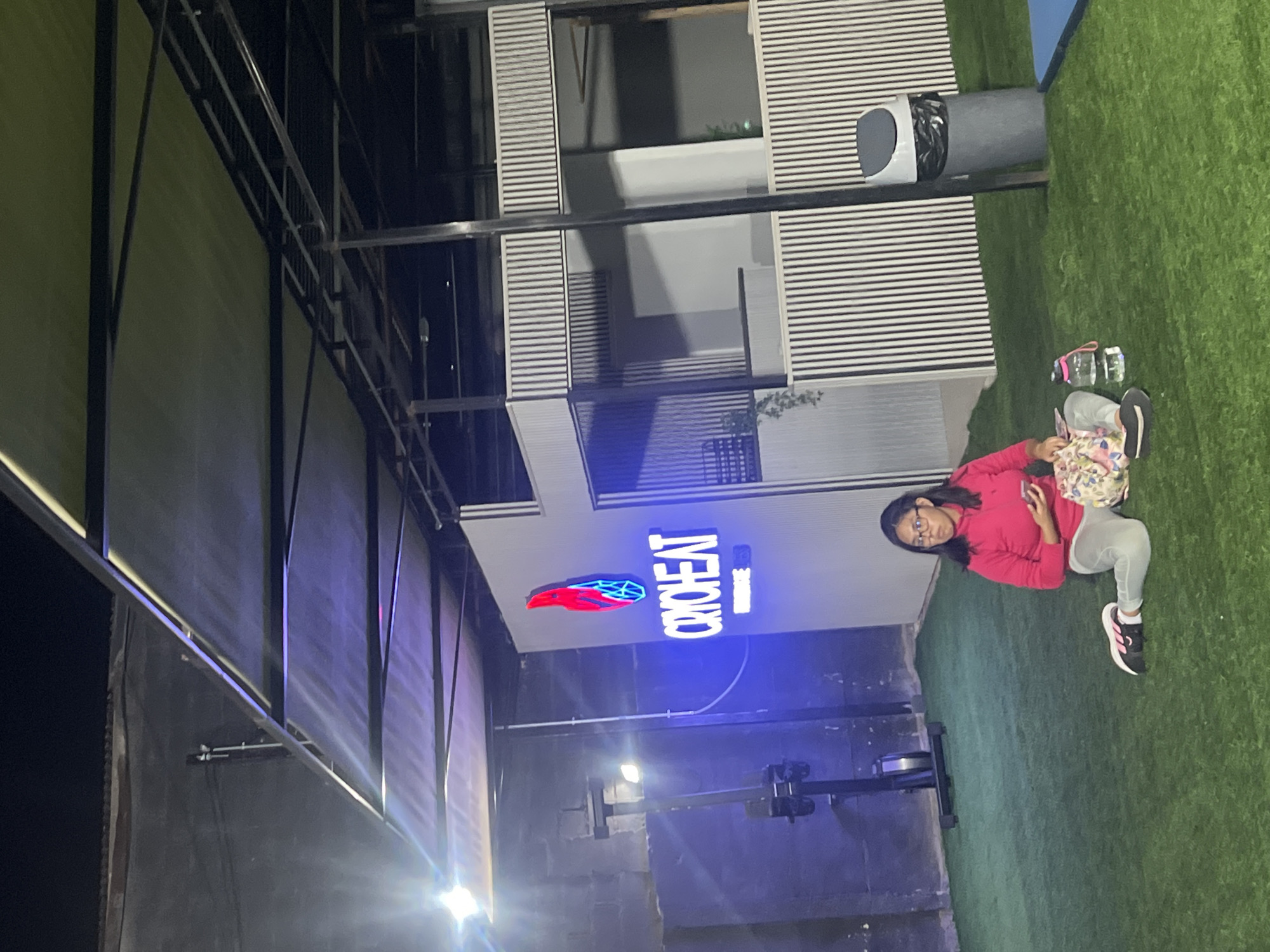 The image shows a woman seated on artificial turf near a building with a sign that reads "Cryoheat." The building is modern with horizontal lines and a blue neon sign. There's also an indoor rowing machine nearby. The location appears to be in an industrial or commercial area, possibly in Arica, Chile, as indicated by the prompt. The woman is wearing a pink sweatshirt, light gray pants, and black and pink sneakers. She is holding a phone and sitting on a patterned bag. A trash can is positioned near the building's edge, and structural elements of the building are visible overhead. The lighting is mixed, with natural light and the neon glow of the Cryoheat sign.