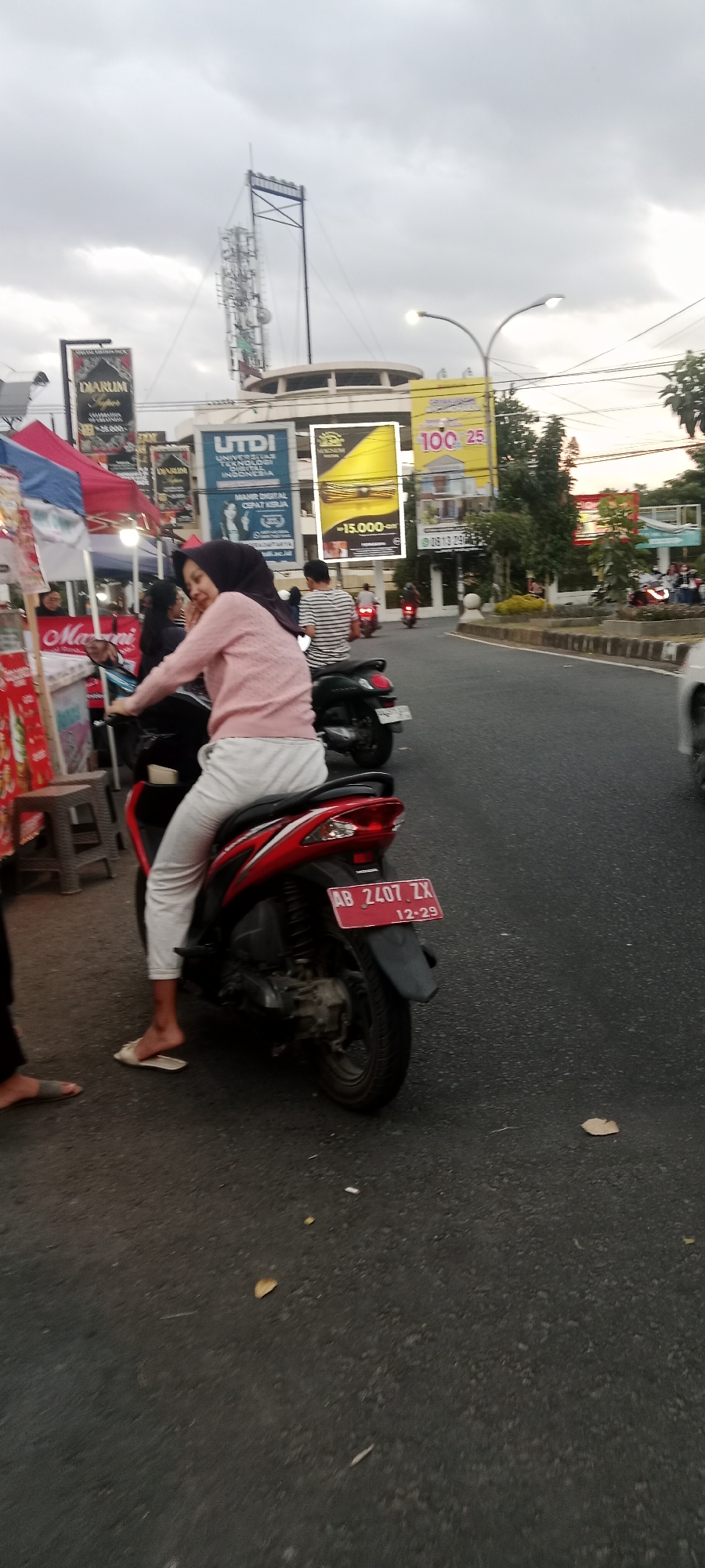 In the city of Ngemplak, Indonesia, a woman wearing a pink sweater and a dark hijab is riding a red and black motorcycle. She is looking over her shoulder, smiling. The motorcycle's license plate reads "AB 2407 ZX 12-29". In the background, there are several billboards and traffic lights. The sky is overcast, suggesting it is late afternoon or early evening. The road is paved and there are other vehicles and pedestrians visible in the distance.