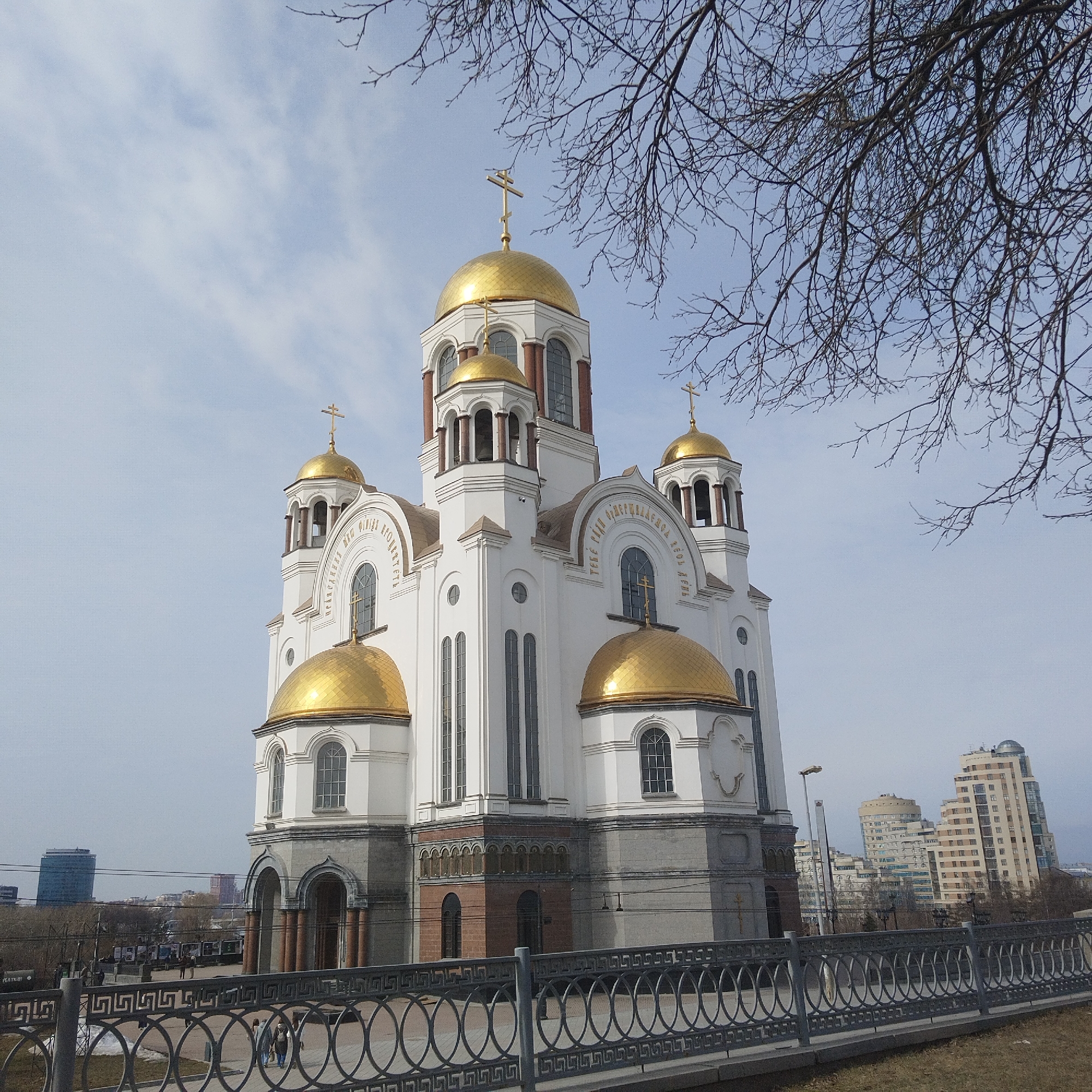 The image shows the Church on Blood in Yekaterinburg, Russia, a large white Orthodox church with golden domes. The building is situated behind a gray metal fence. Several high-rise buildings can be seen in the background. Some bare tree branches occupy the upper right corner of the frame. People are walking on the plaza in front of the church. The sky is overcast.