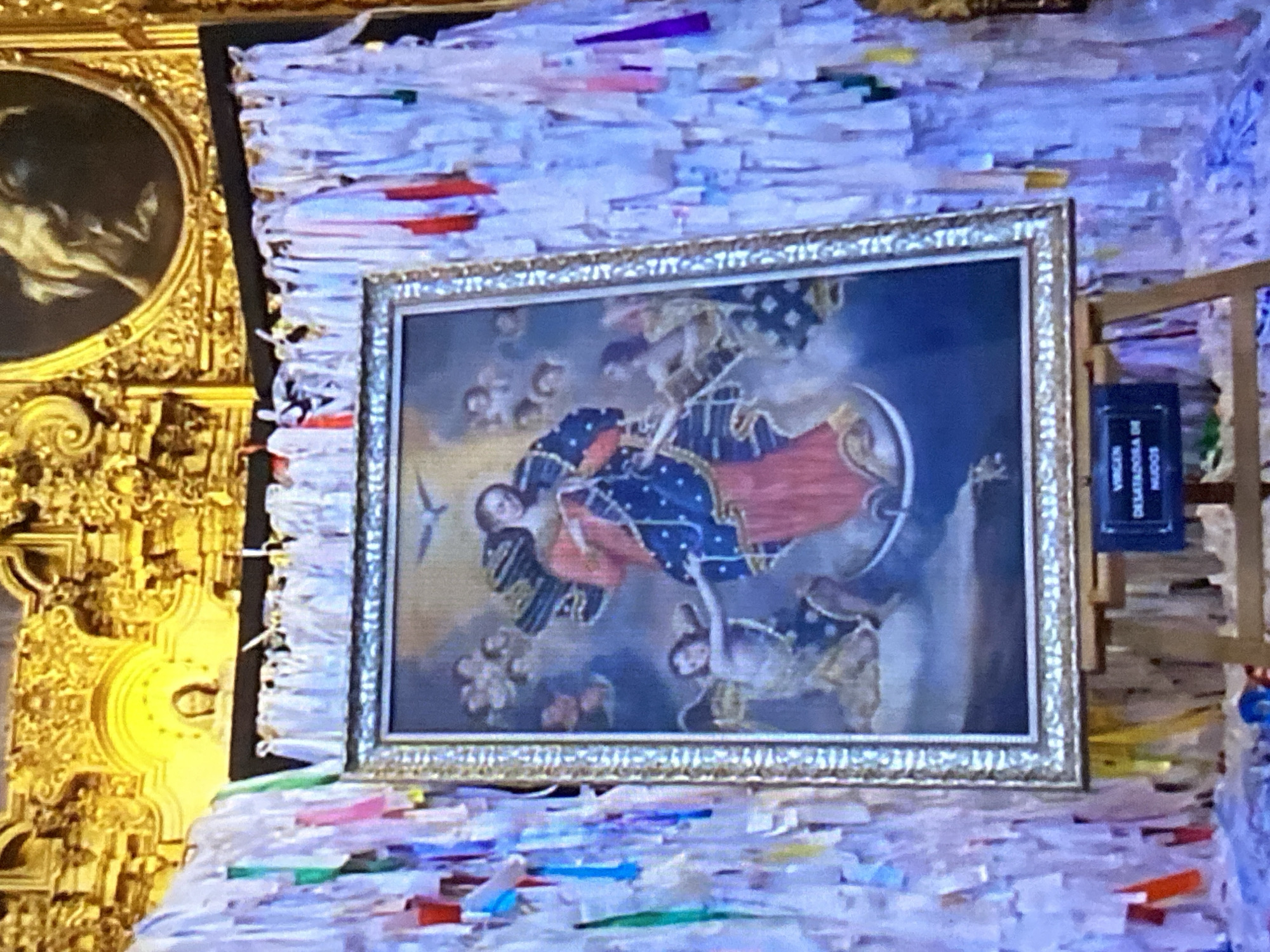 The image shows a framed painting depicting the Virgin Mary, likely Our Lady of the Knots (Virgen Desatadora de Nudos). The painting is hung against a wall covered in numerous small white ribbons or strips of paper. The lower portion of an ornate gold structure, potentially part of an altar or architectural detail, is visible on the left side of the image. To the right of the painting is a small, dark sign with white lettering that says "VIRGEN DESATADORA DE NUDOS." The location is likely in Tinaquillo, Venezuela, given the probable subject matter and the presence of the Spanish text on the sign. The scene appears to be a place of devotion or pilgrimage.